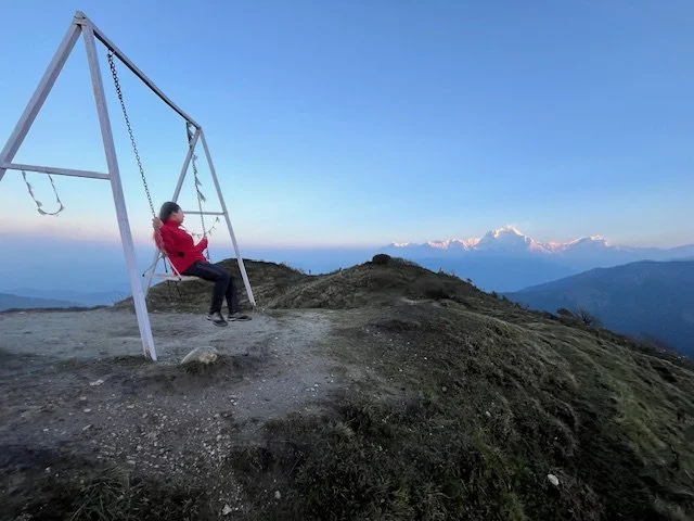 A person sitting on a swing on a mountain overlook with a view of distant mountains and clouds.