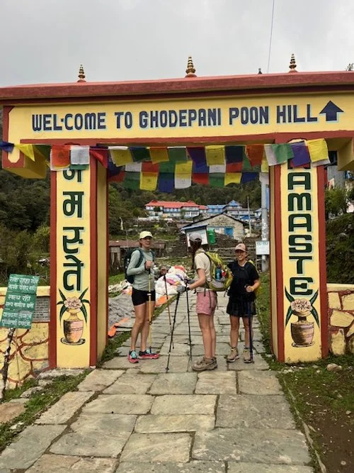 Trekkers in hiking gear standing under a colorful welcome arch at Ghodepani Poon Hill in Nepal, with village houses and mountains visible in the background.