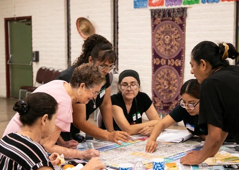 Group of women gathered around a table, talking together about their neighborhood and their families, inside a room a community learning room.