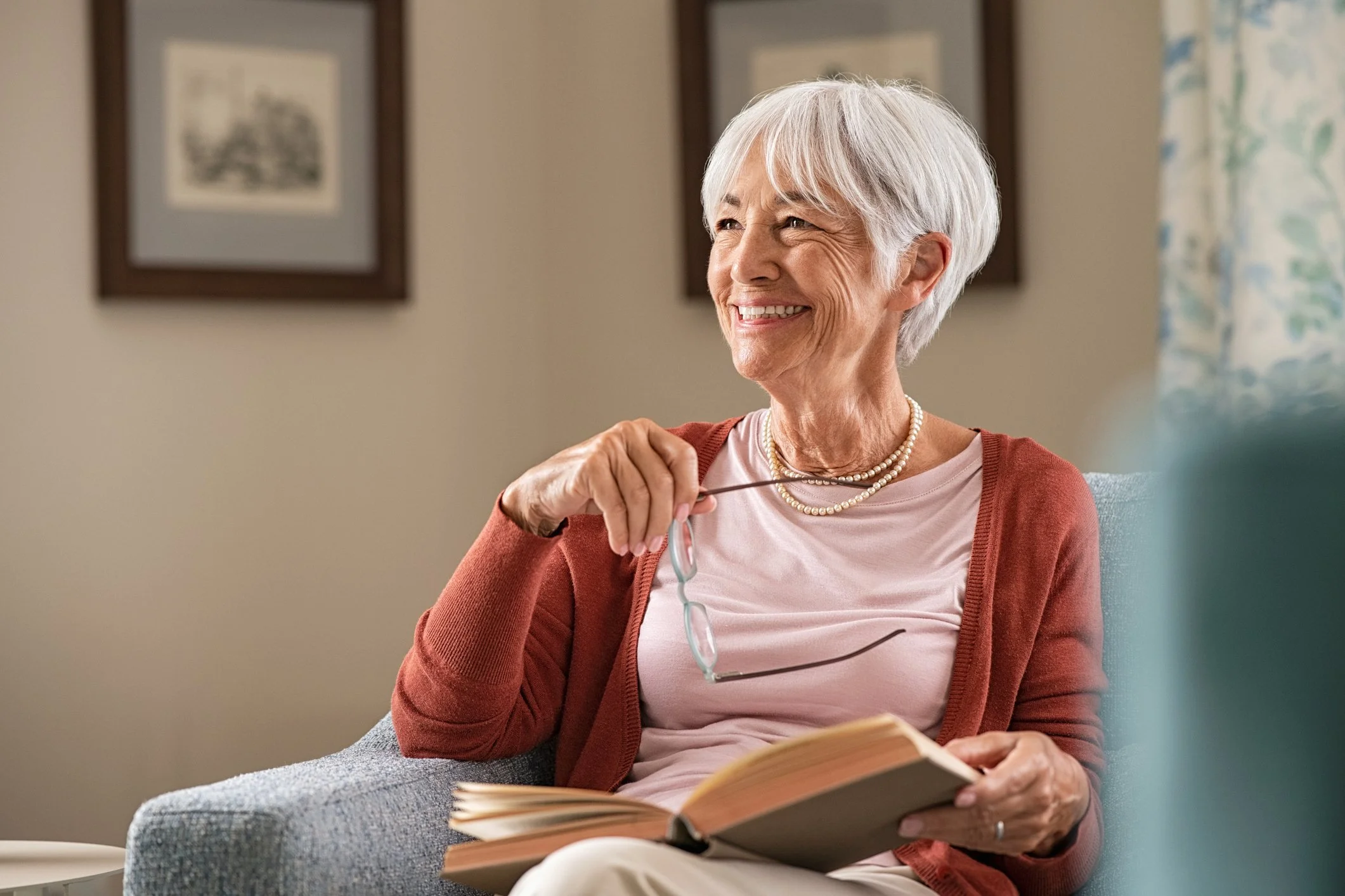 An elderly woman with white hair smiling while sitting on a couch, holding reading glasses and a book, with framed pictures and a floral curtain in the background.