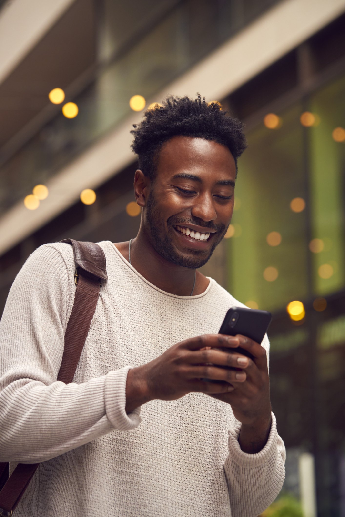 A young African American man smiling and looking at his phone outside in an urban setting at night with blurred lights in the background.