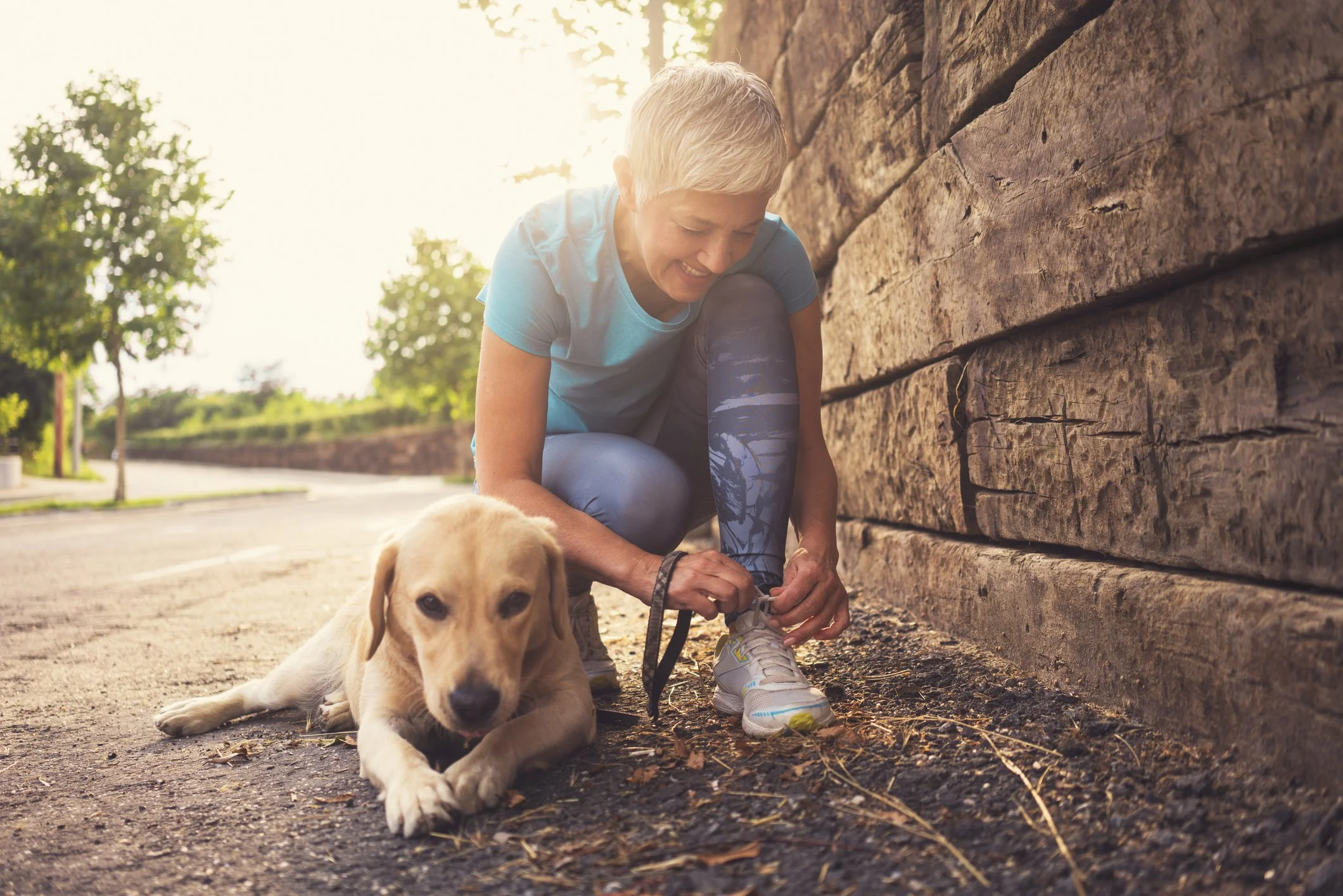 A woman with short blonde hair in athletic clothing crouches down to put a leash on her tan puppy, sitting on a sidewalk near a wooden wall, with greenery and trees in the background.