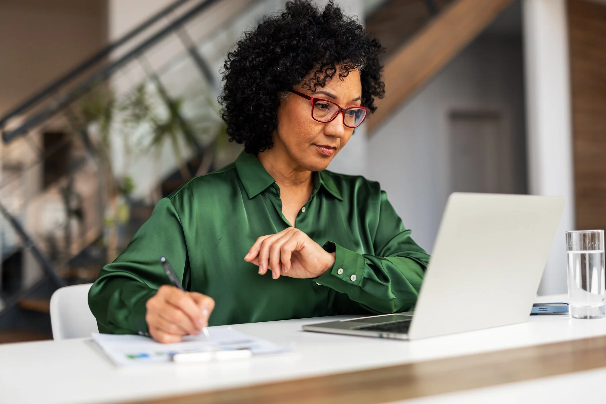 A professional woman with curly black hair, red glasses, and a green satin blouse sitting at a white desk with a laptop and glass of water, in a modern workspace.