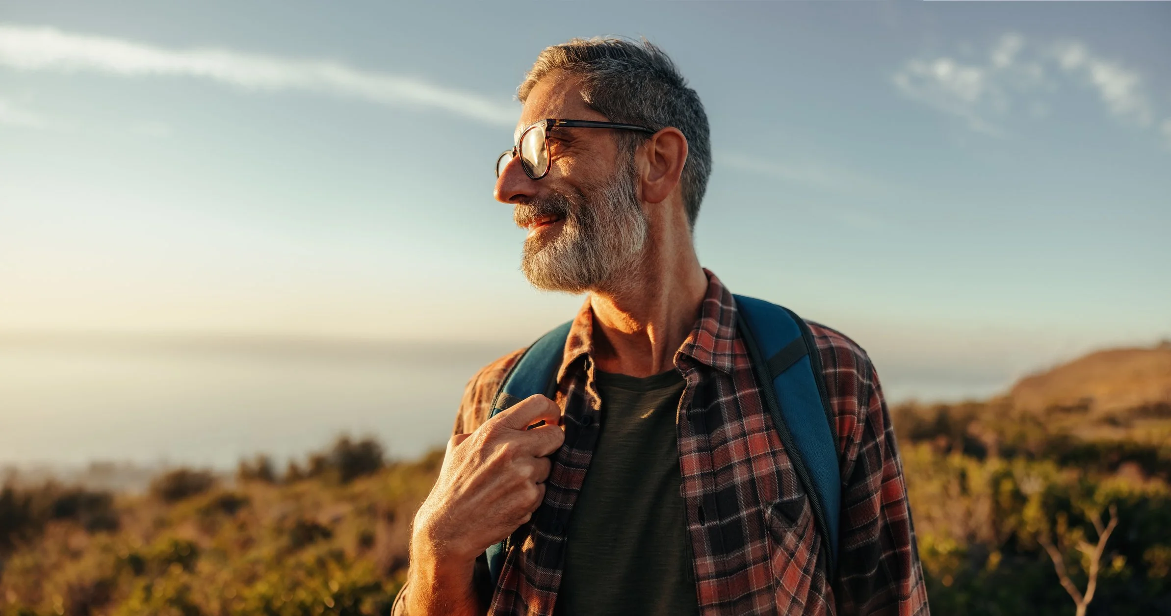 A smiling man with gray hair, beard, and sunglasses standing outdoors during sunset, wearing a plaid shirt and carrying a backpack.