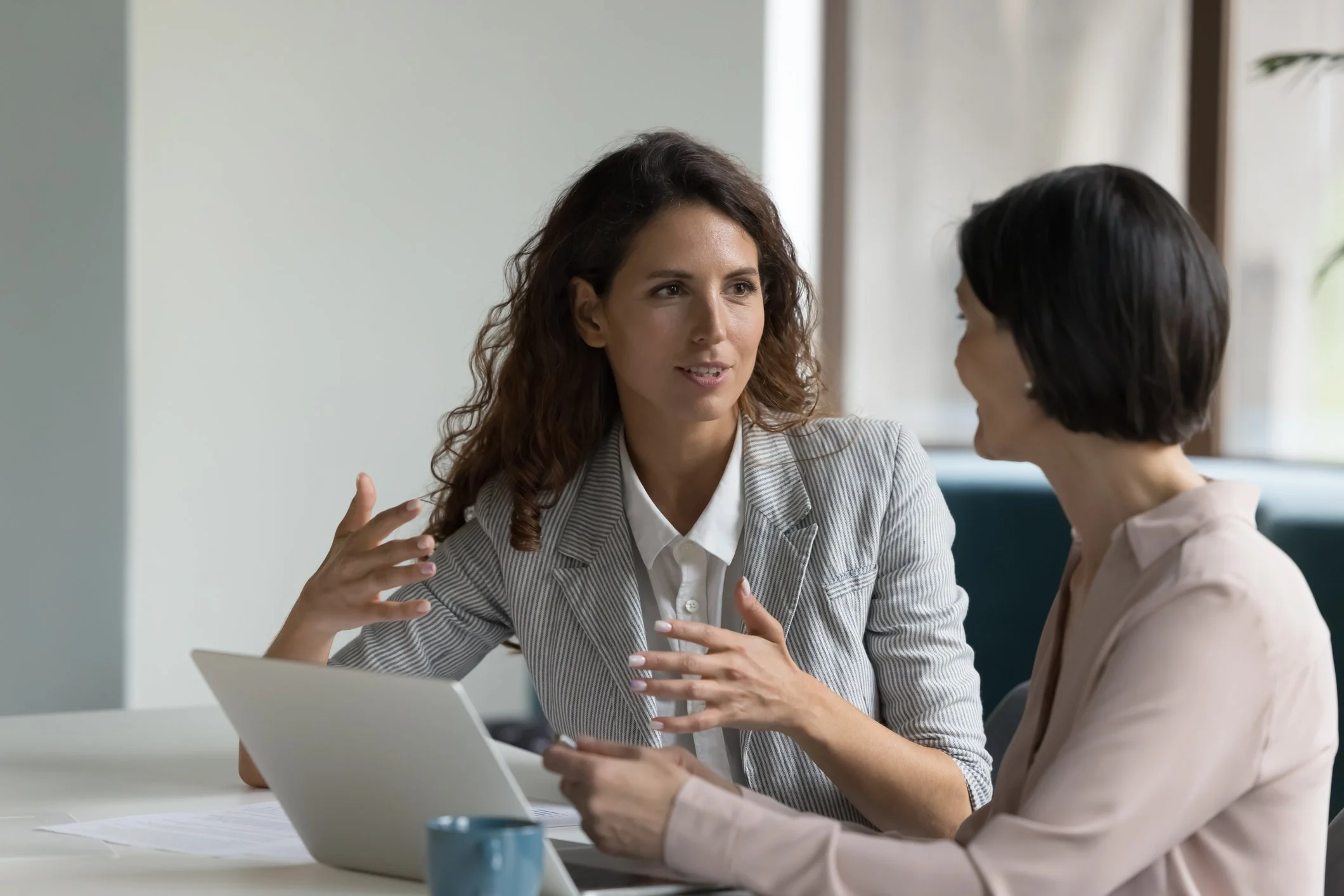 Two women professionals engaged in a discussion at a desk with a laptop, papers, and a coffee mug on the table.