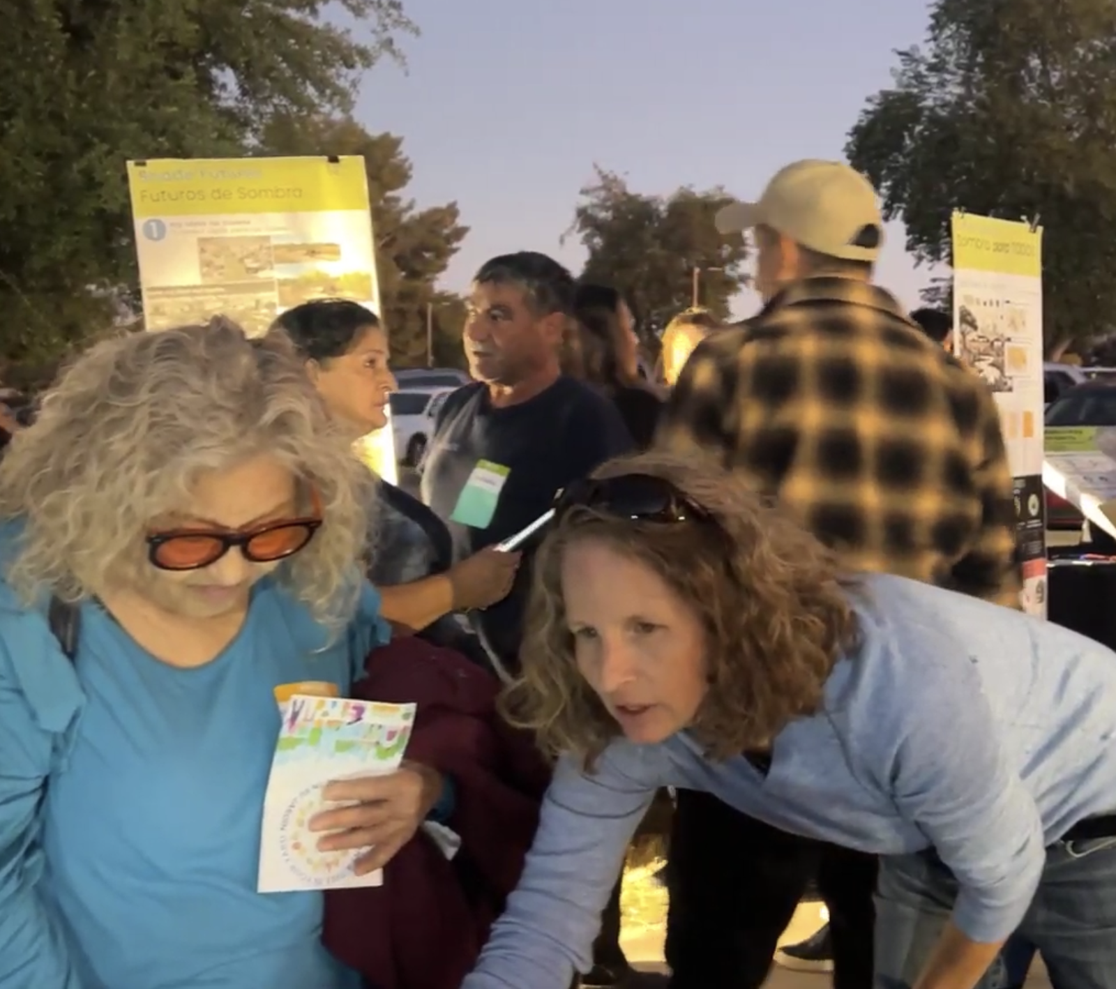 Group of people gathered outdoors during the evening with informational posters in the background, possibly at an event or gathering.