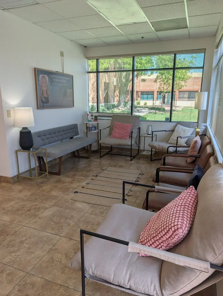 A waiting room at Jen Clifton Counseling & Psychotherapy with multiple chairs and a bench, two lamps, a display screen on the wall, and large windows showing trees and a building outside.