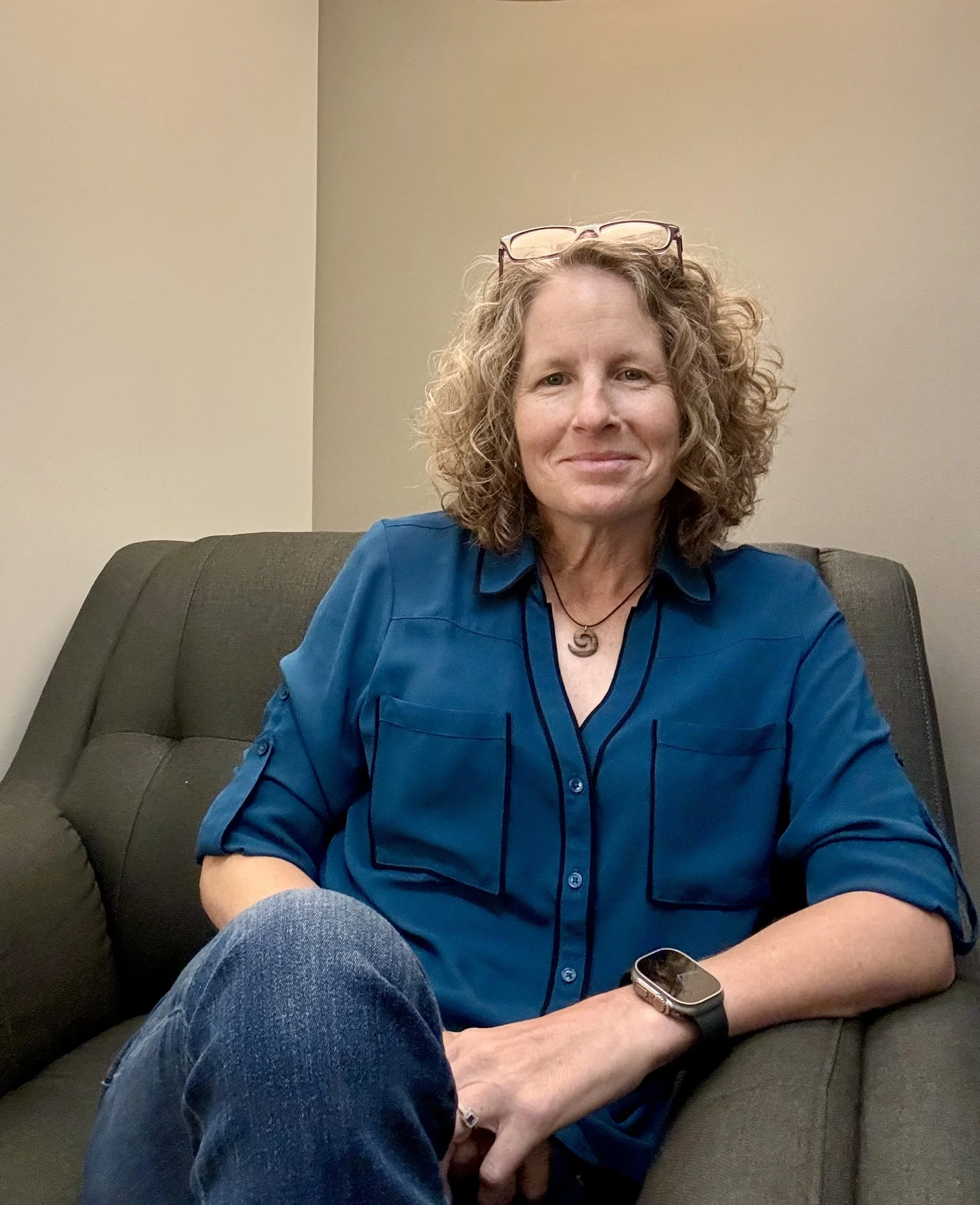 Jen Clifton, a therapist in Arizona, with curly hair, sitting on a dark gray sofa, wearing a blue shirt and a smartwatch, with glasses resting on her head, in a therapy room in Gilbert, Arizona.
