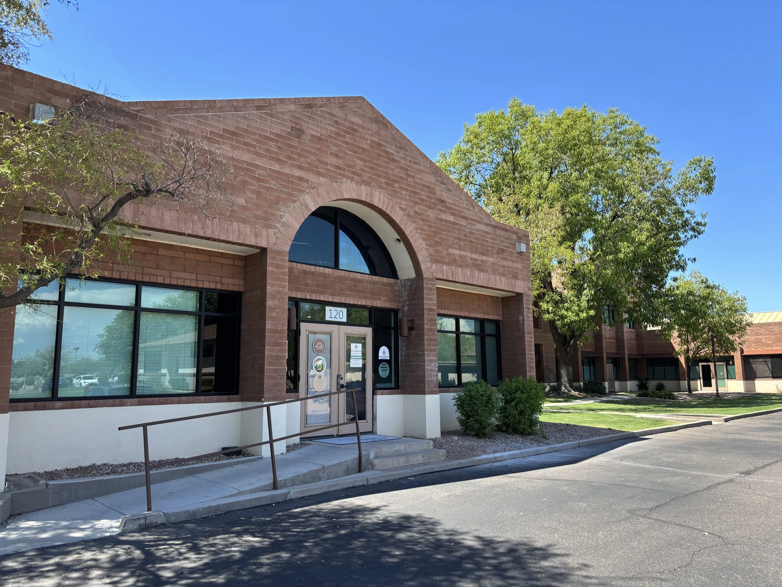 Jen Clifton Counseling & Psychotherapy office building. Red brick building with large arched windows, trees and a sidewalk in front, and a clear blue sky.
