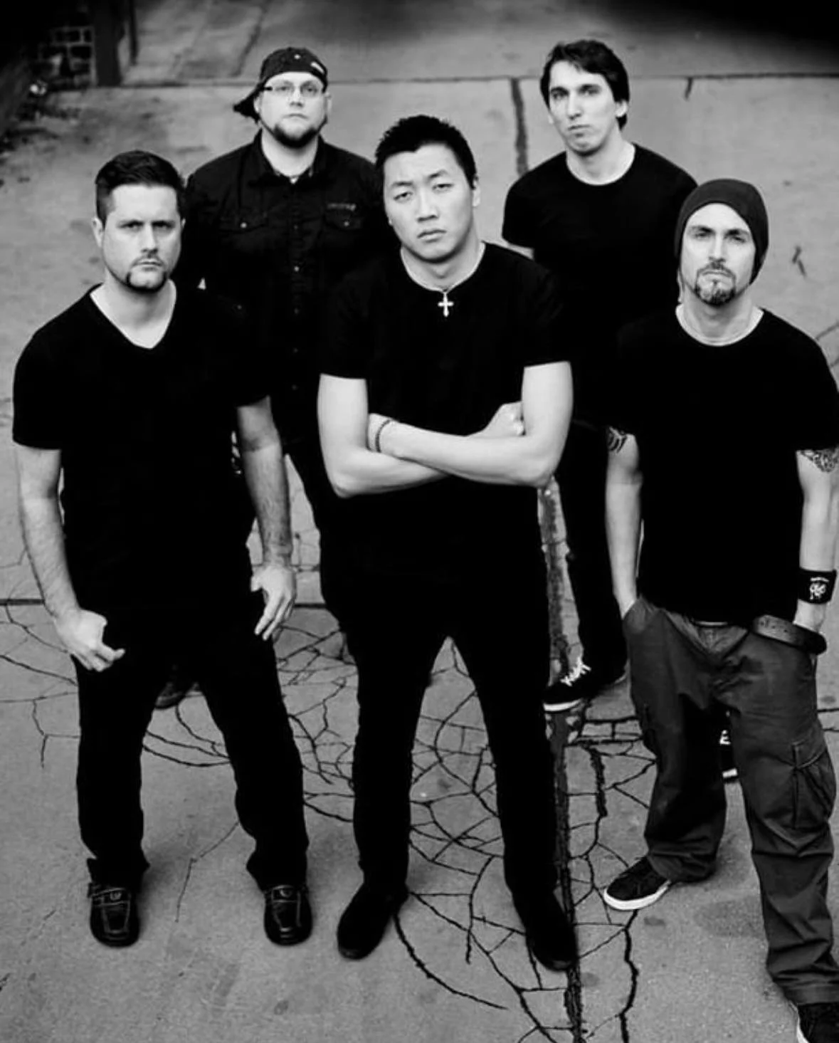 Black and white photo of six men standing on a cracked pavement, with one man in front has his arms crossed.