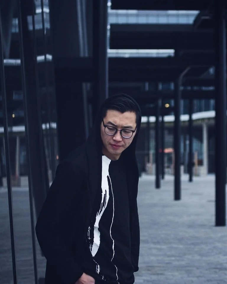 A young man wearing glasses, a black hoodie, and a black jacket, standing outdoors near a modern glass building, looking down with a serious expression.