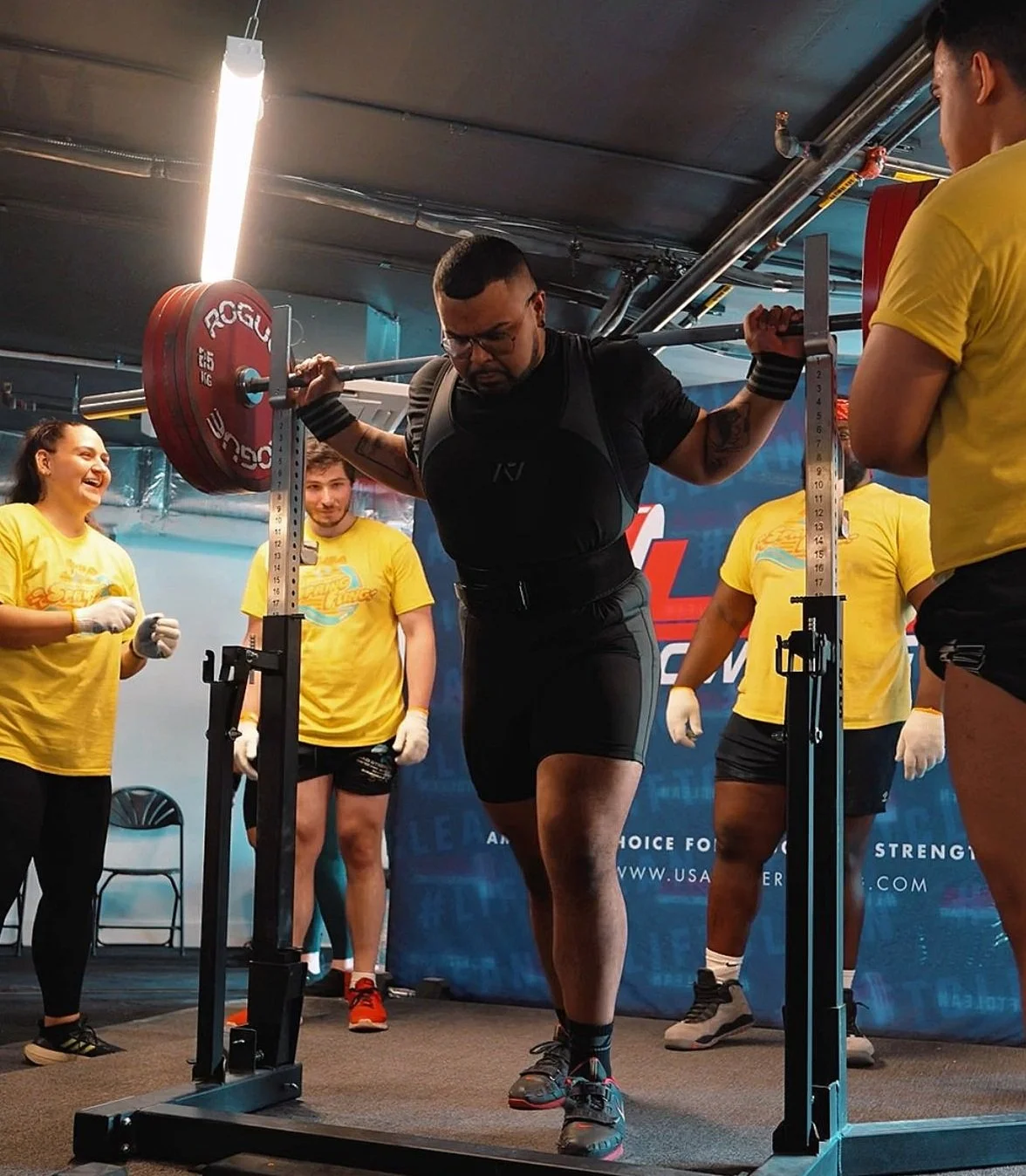 Man in black athletic gear performing a squat with a barbell in a gym setting with onlookers in yellow shirts