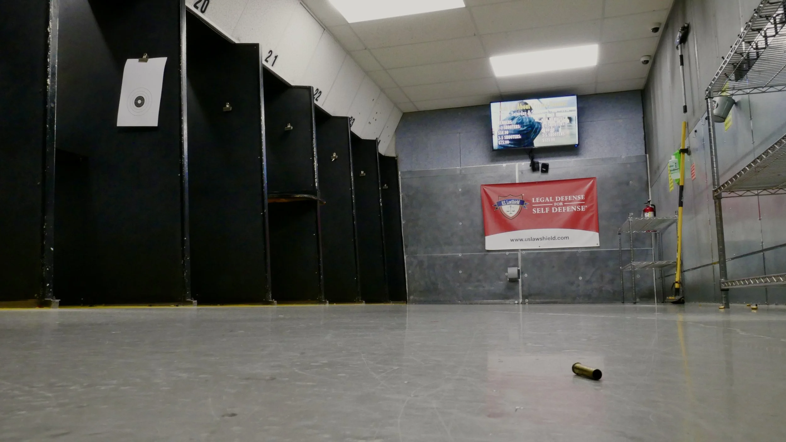 Empty indoor shooting range with black shooting bays, TV monitor, red banner, and empty shelves. Bullet casing on floor.