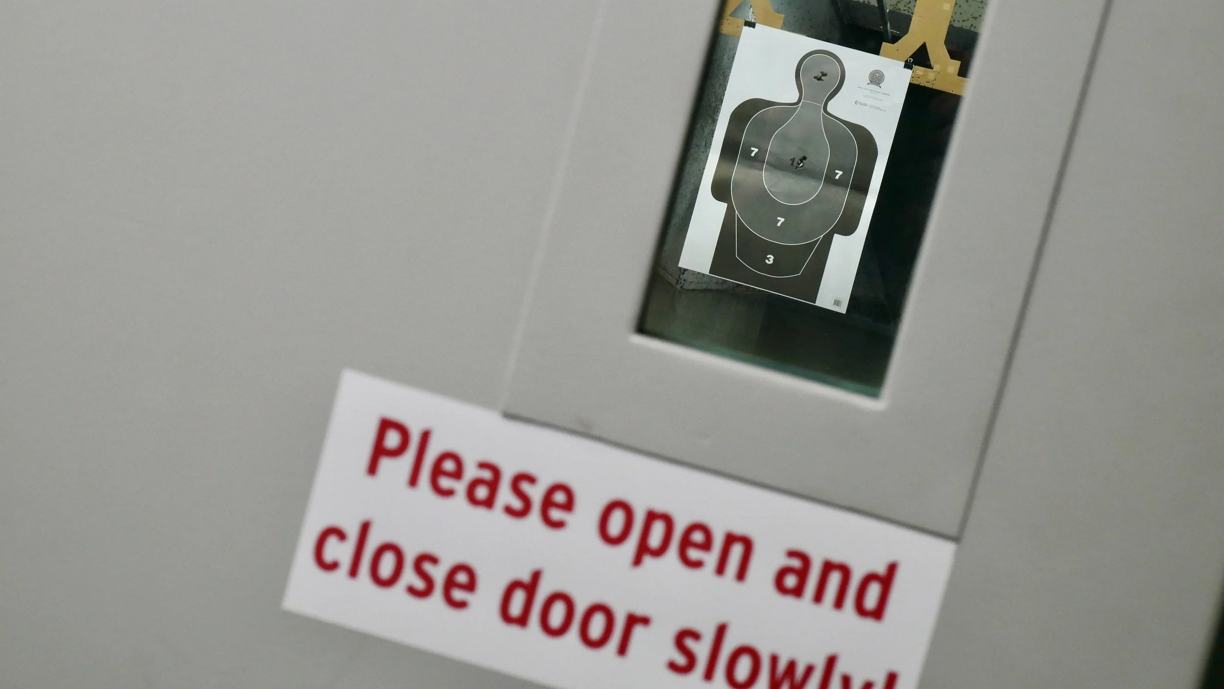 A private shooting bay booth door that reads "please open and close door slowly", through the window you can see a target with tight grouping to the head and chest.