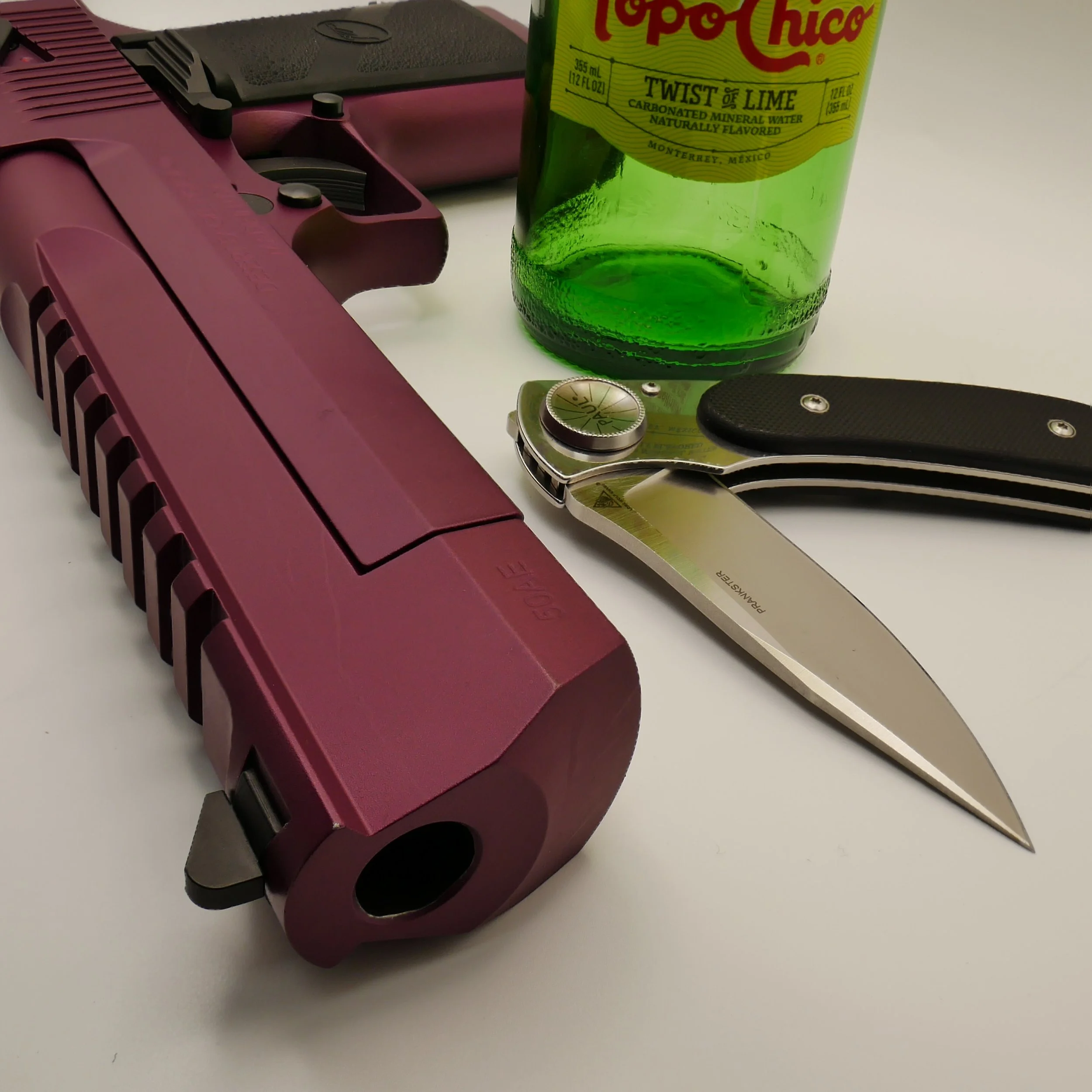 A plum Desert Eagle .50 Caliber pistol, is next to a green bottle of Topo Chico Twist of Lime sparkling water, and a Chris Reeve's Ikosi pocket knife with a Ebony handle and partially open blade on a white surface.