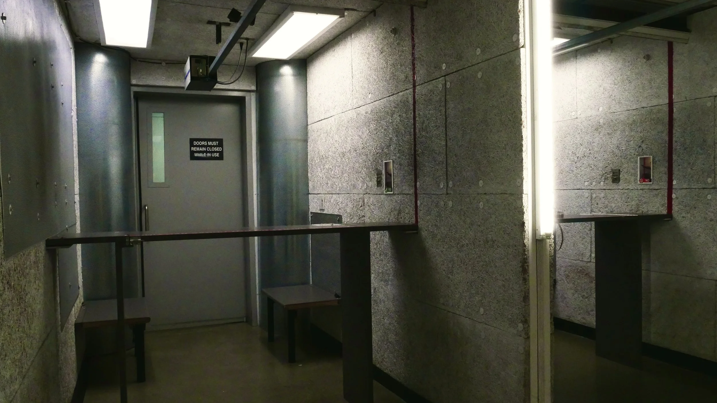 A small lit private shooting booth with gray sound proof walls. There is a single counter with a bench underneath on the left side. Behind a gray door with a window, there is a sign that reads 'Doors must remain closed while in use.'