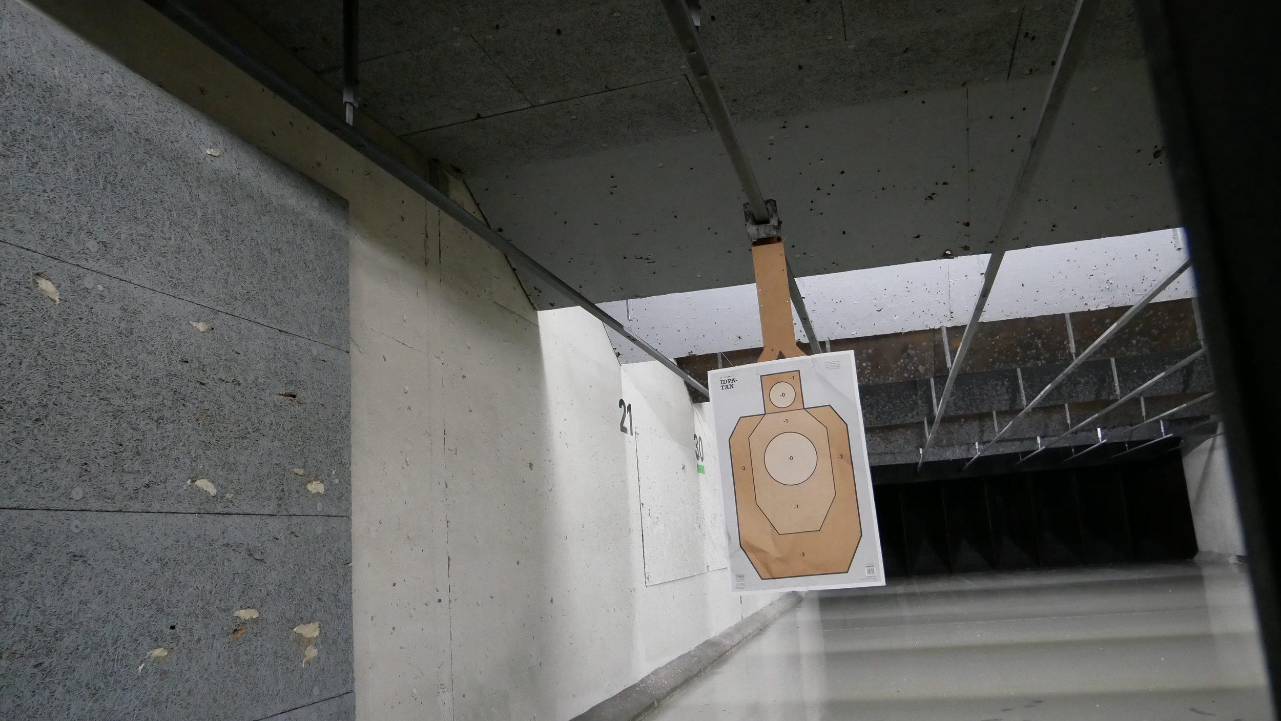 Indoor shooting range with target sheets, including a paper target with concentric circles, hanging from a hook on a metal support, with numbered walls and a dark background.