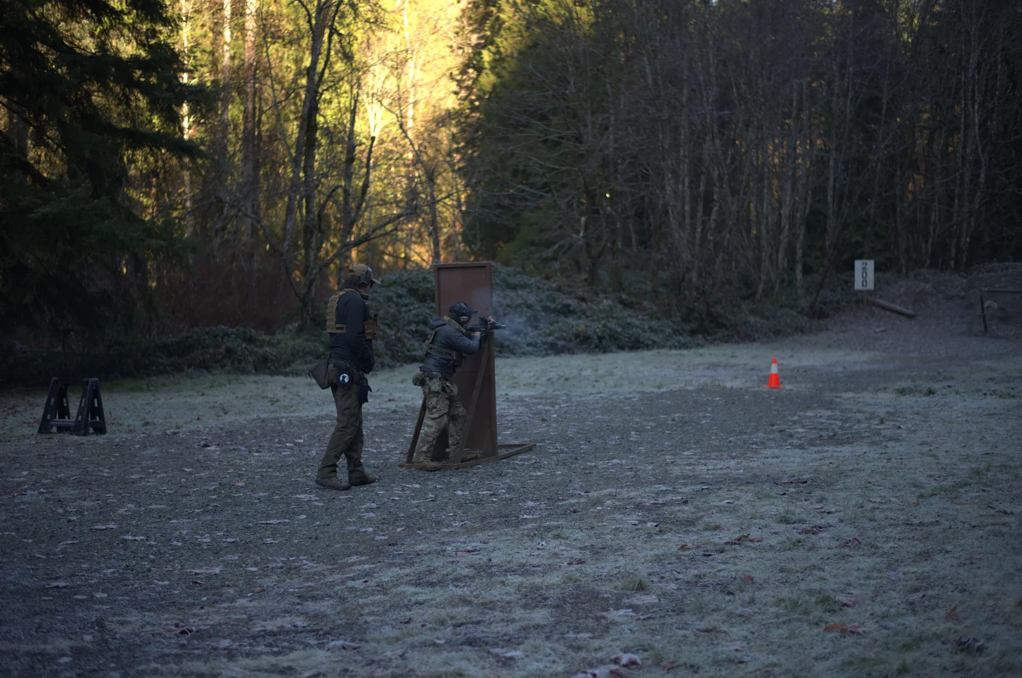 Two tactical officers practicing firearms training outdoors on a frosty morning, one aiming a gun at a target on a shooting range, in a wooded area with trees and orange traffic cone in the background.