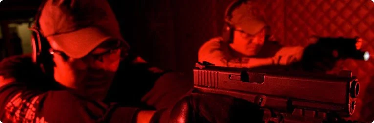 Two individuals wearing protective glasses and headphones, aiming guns in a dimly lit, red-colored environment.