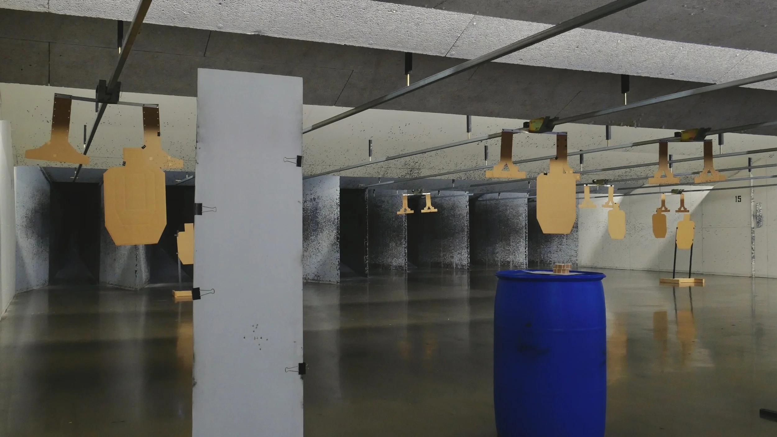 Indoor shooting range with UPSCA/IPSC cardboard targets hanging on stands and a blue barrel on the floor making a barricade. The targets are arranged in a course of fire set for competitive use.