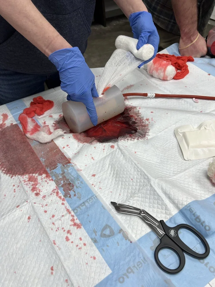 Person wearing blue gloves handling a tissue swab over a blood spill on medical pad, with medical tools and supplies nearby.