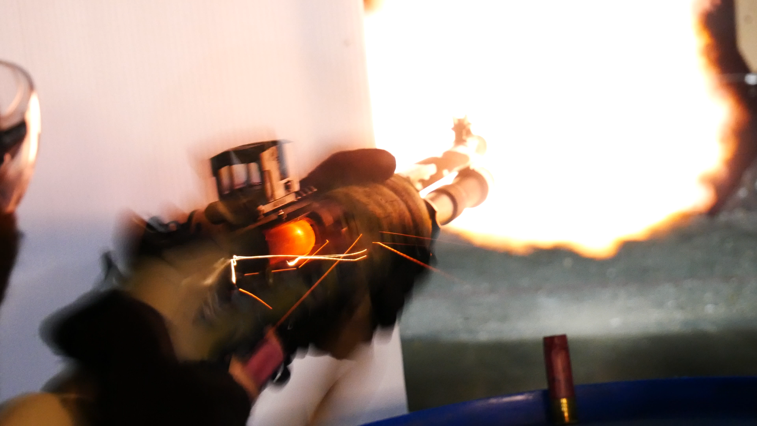 A person firing a benelli m4 clone called the MAC1014 shotgun, with a visible large flame and sparks coming from the barrel. He is shooting through a barricade at the indoor range.