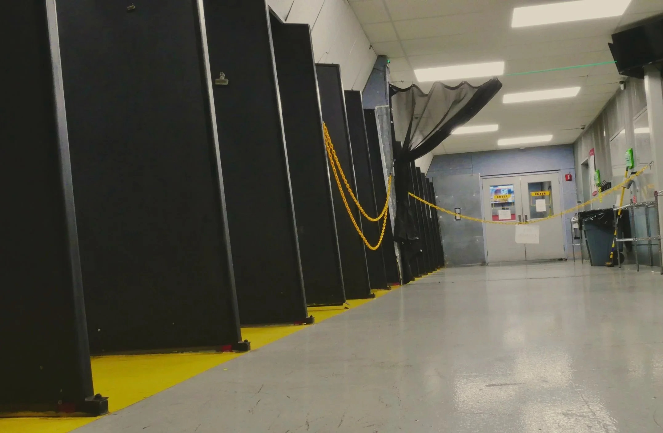 Empty indoor shooting range with black shooting bays, a door in the back,  grey floor, red banner, and empty shelves. Bullet casing on floor.