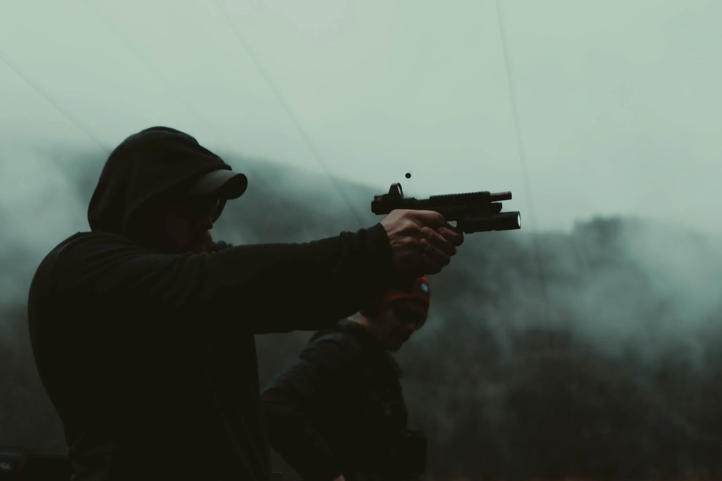 Person in dark hoodie aiming a handgun outdoors on a cloudy day.