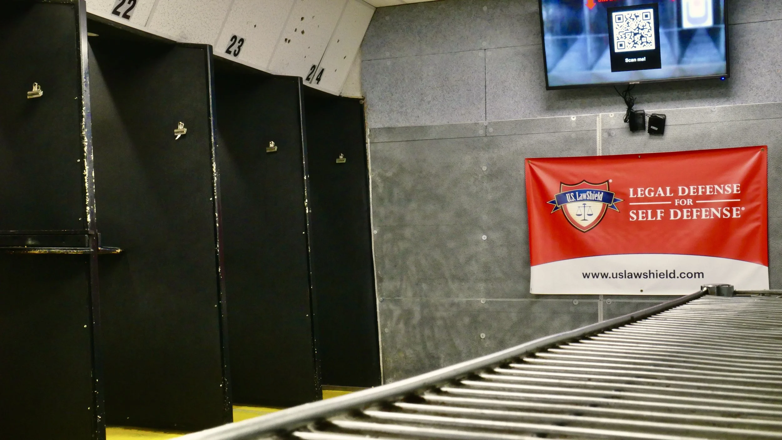 Empty indoor shooting range with black shooting bays, TV monitor showing a qr code, red banner reading US Law Shield, and empty shelves. Bullet casing on floor.