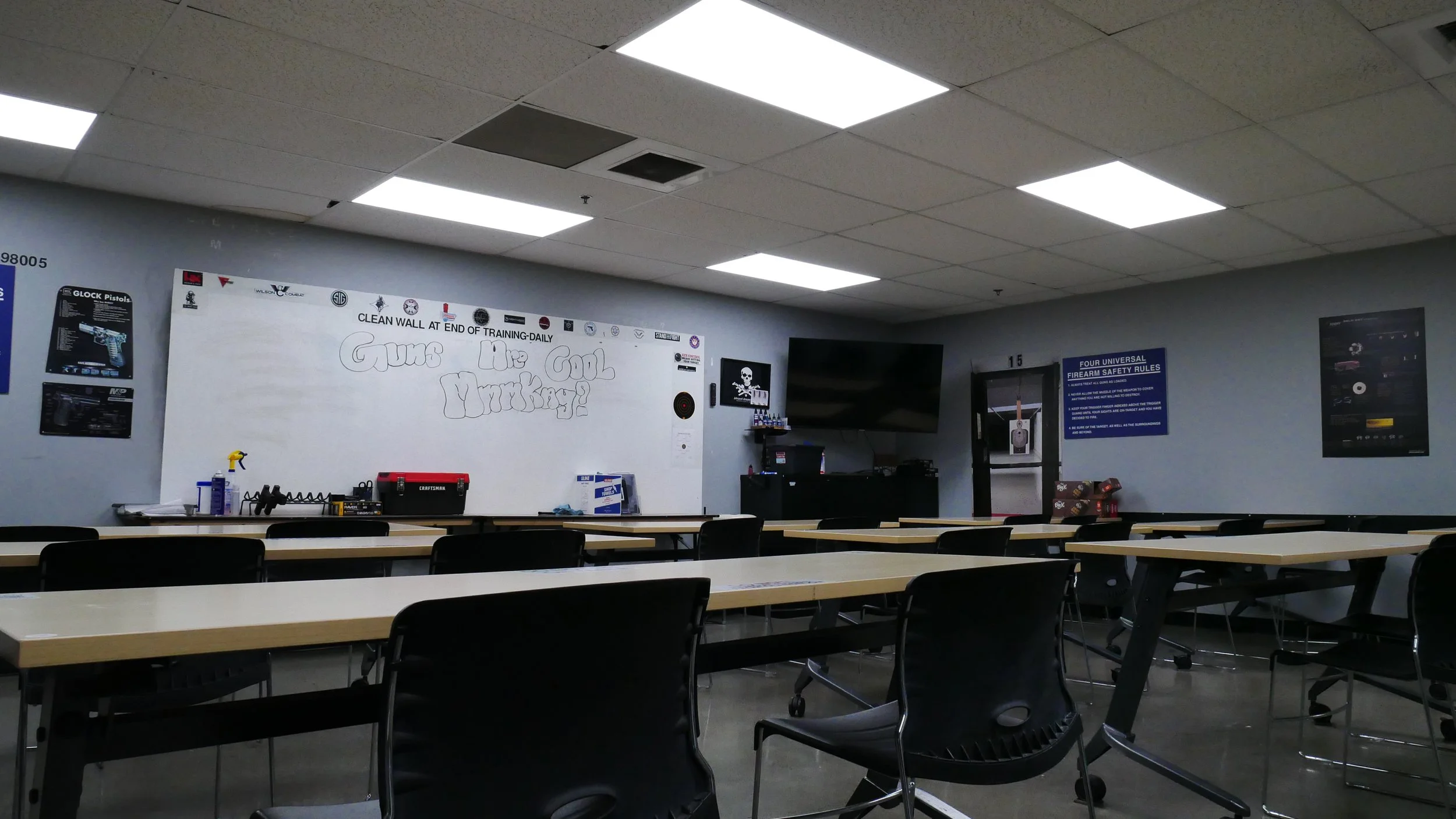 A shooting range classroom with a white board, tv, desks and chairs is prepped for class.