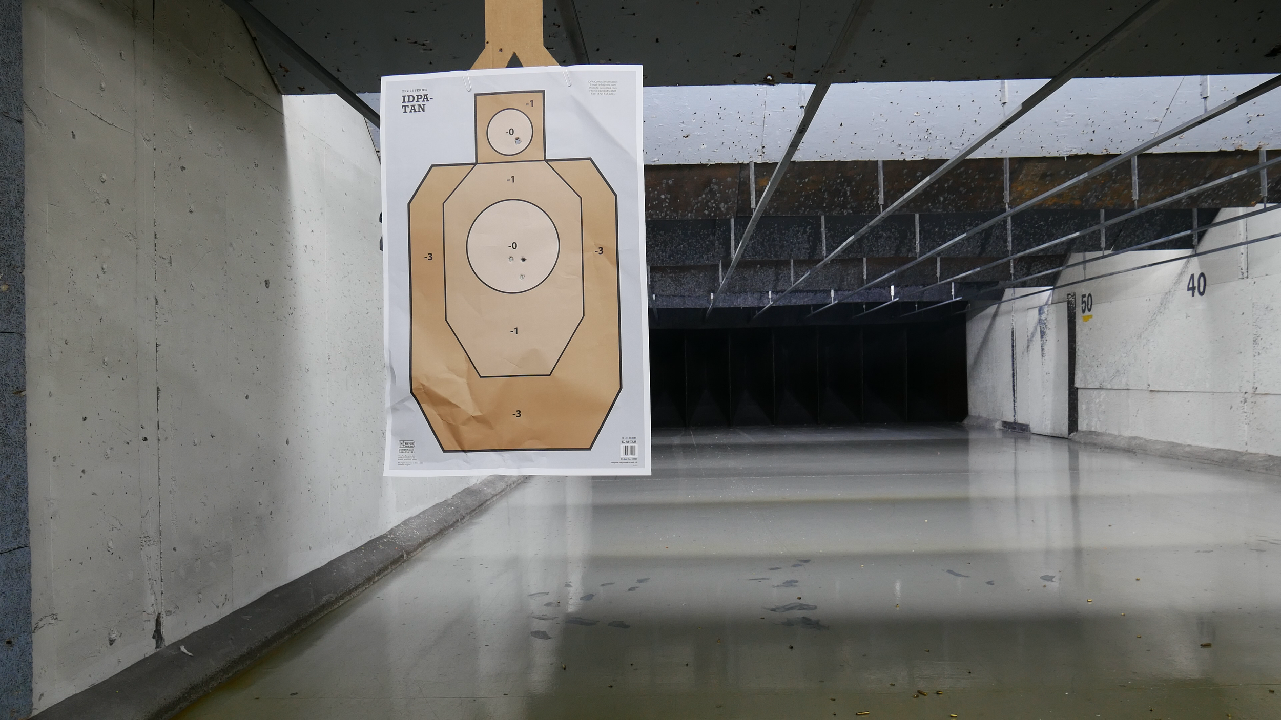 An indoor shooting range with a target paper hanging on the left side. The target has a beige background with black outlines and scoring zones marked for different point values. The concrete floor has some scattered debris, and the ceiling is dark wi