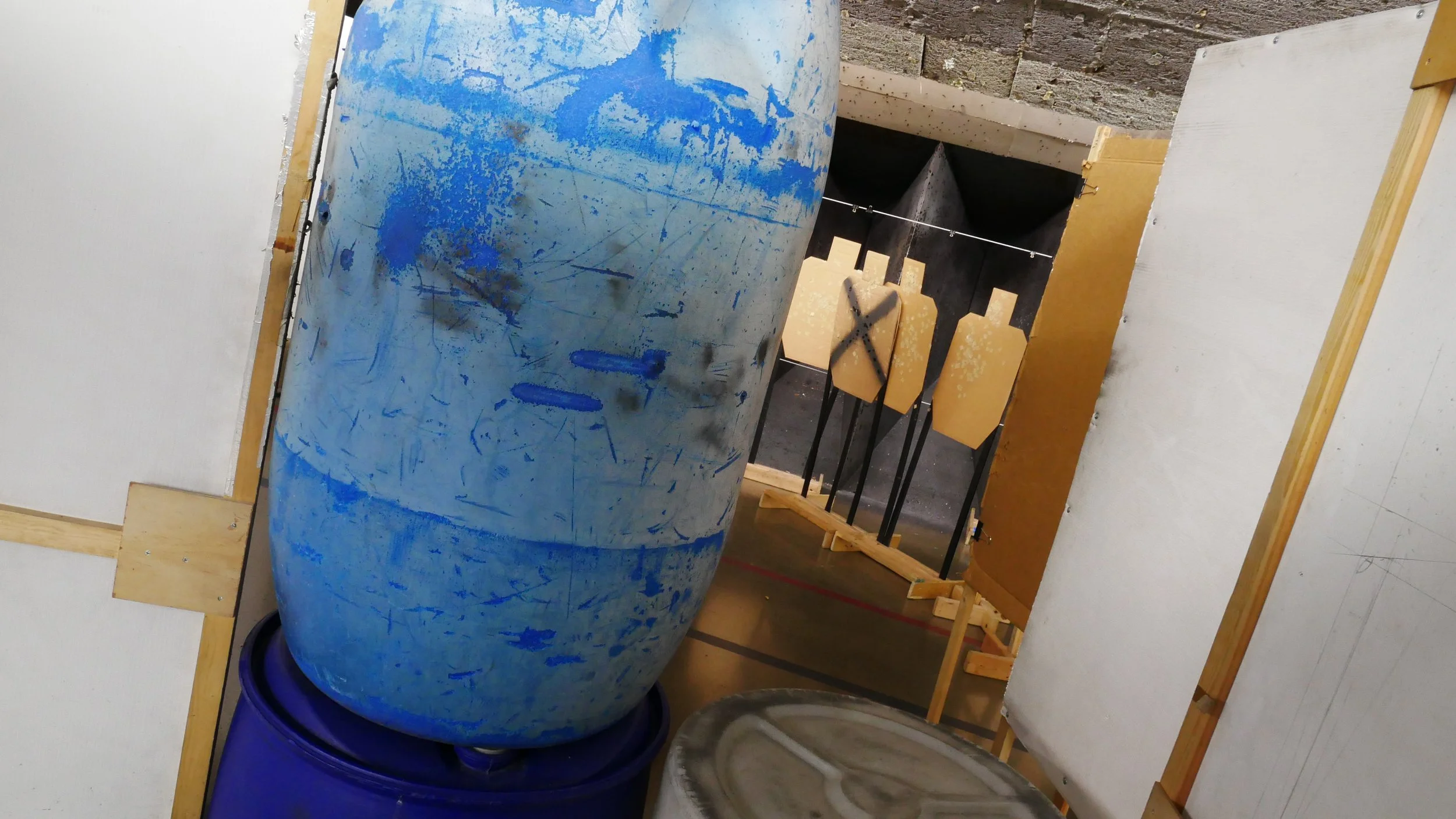 A blue barrel barricade sits between the shooter and cardboard targets at an indoor shooting range.