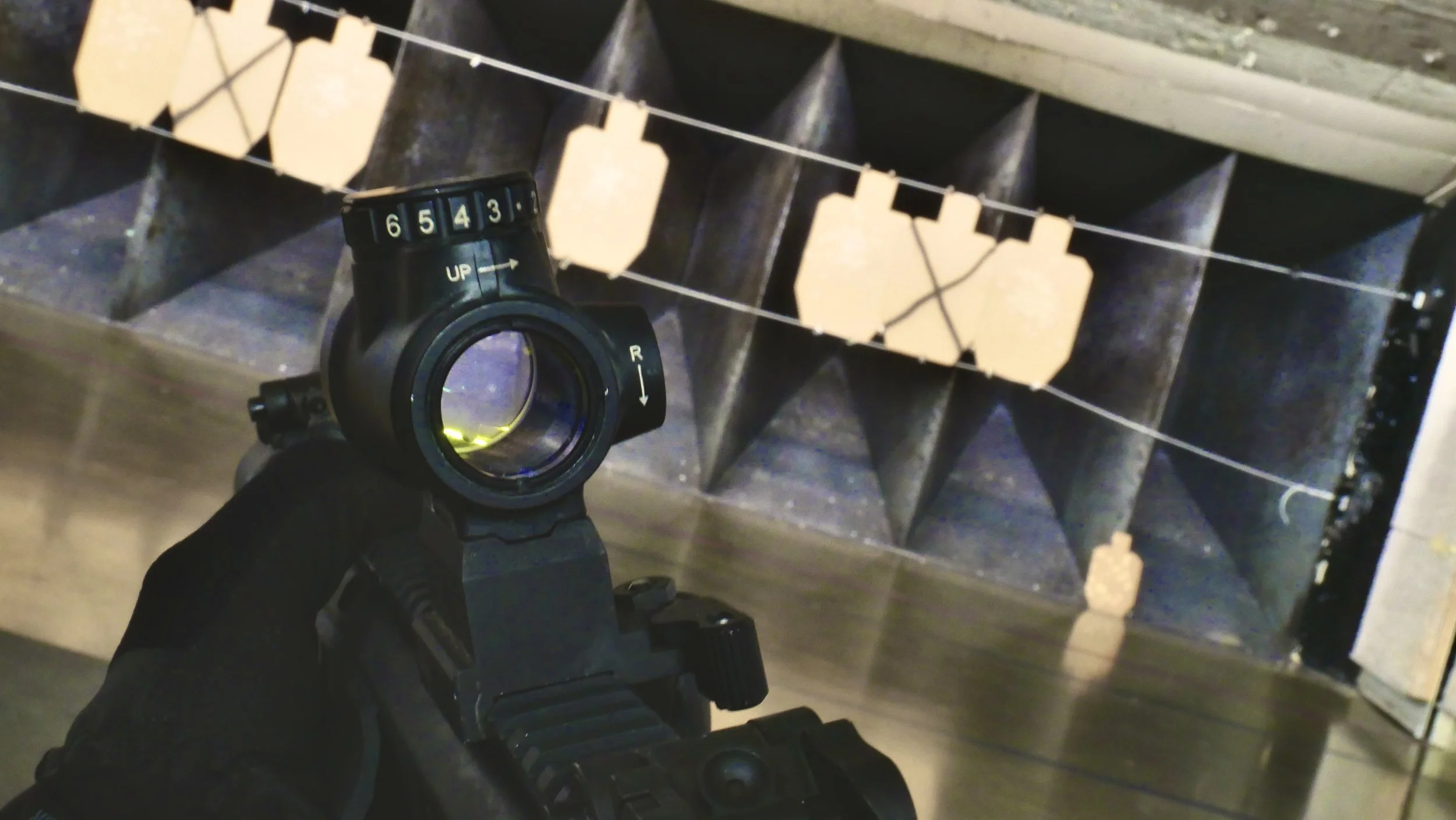 First person view from a person holding a sig sauer mcx with a trijicon mro green dot mounted on top, while he aim down range at targets hanging in an indoor shooting range.