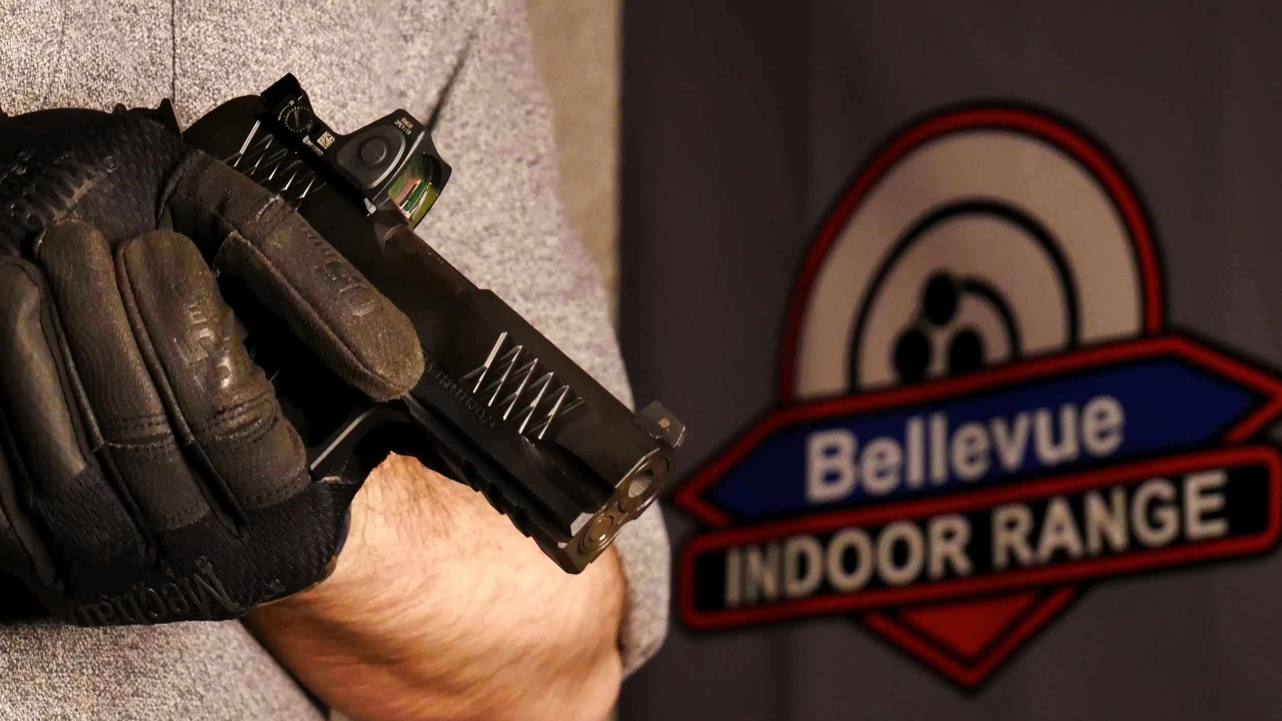 A hand wearing black mechanix gloves holding a black semi-automatic handgun with a green dot sight, near a sign that reads 'Bellevue Indoor Range' in the training bay located in washington.