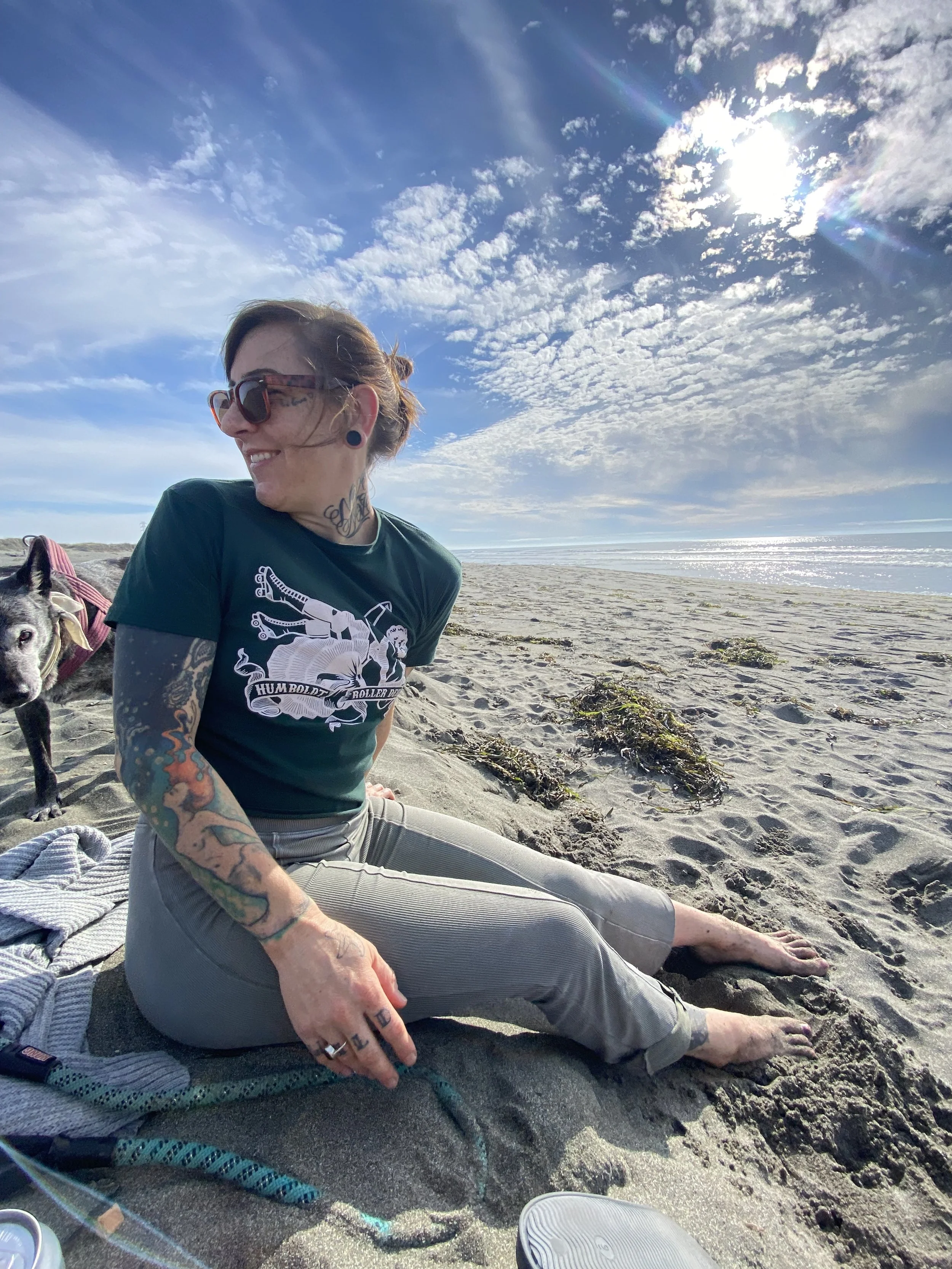 A woman with tattoos and sunglasses sitting on a blanket on a sandy beach, smiling and relaxing as the sun shines brightly in a partly cloudy sky.