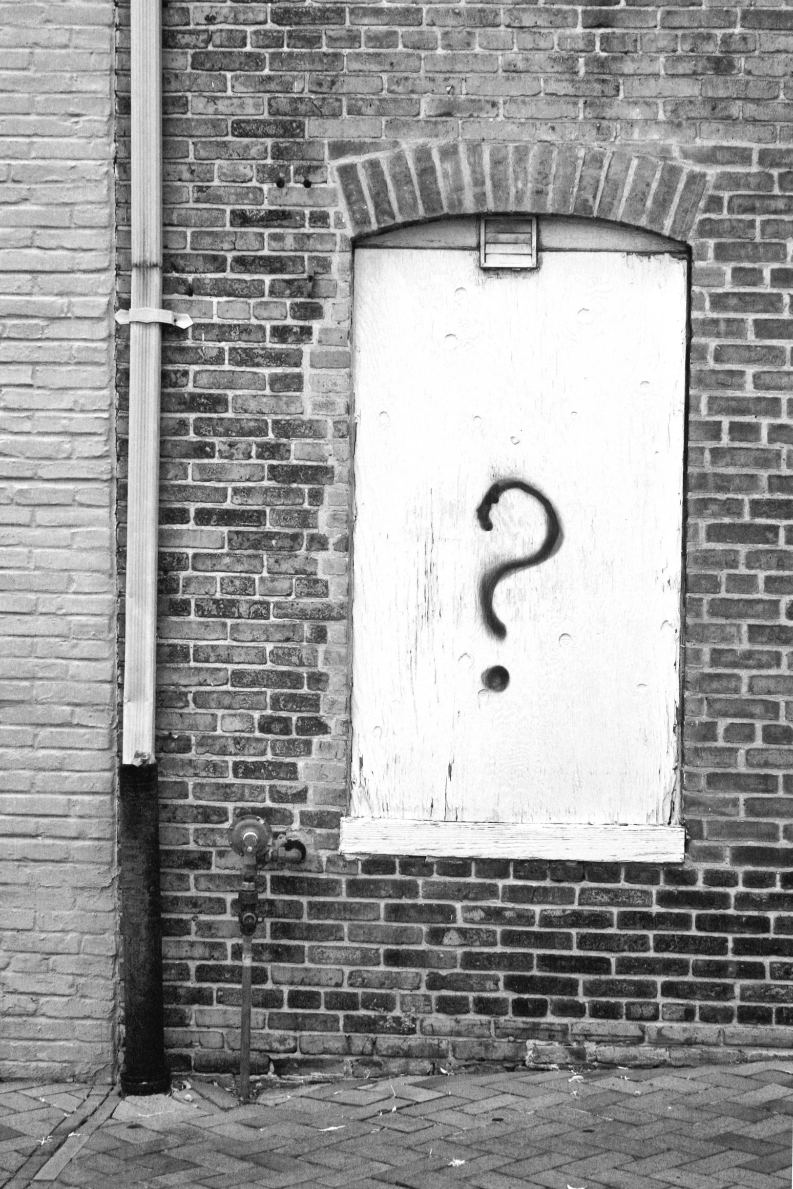 A brick wall with a boarded-up window that has a large spray-painted black question mark on it.