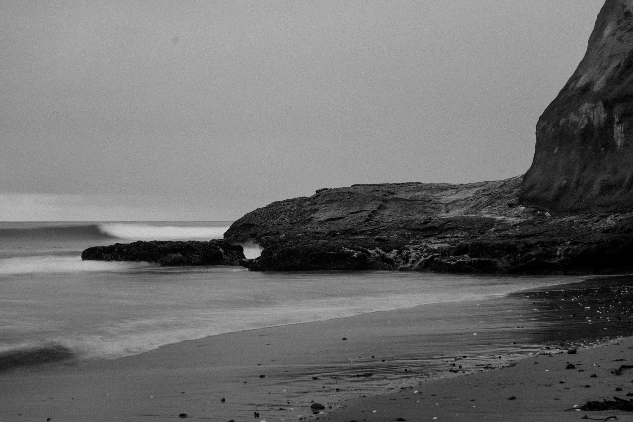 Black and white photo of a rocky shoreline with waves gently hitting the beach, and a large cliff on the right side in the distance. Photo by Vincent Prograce