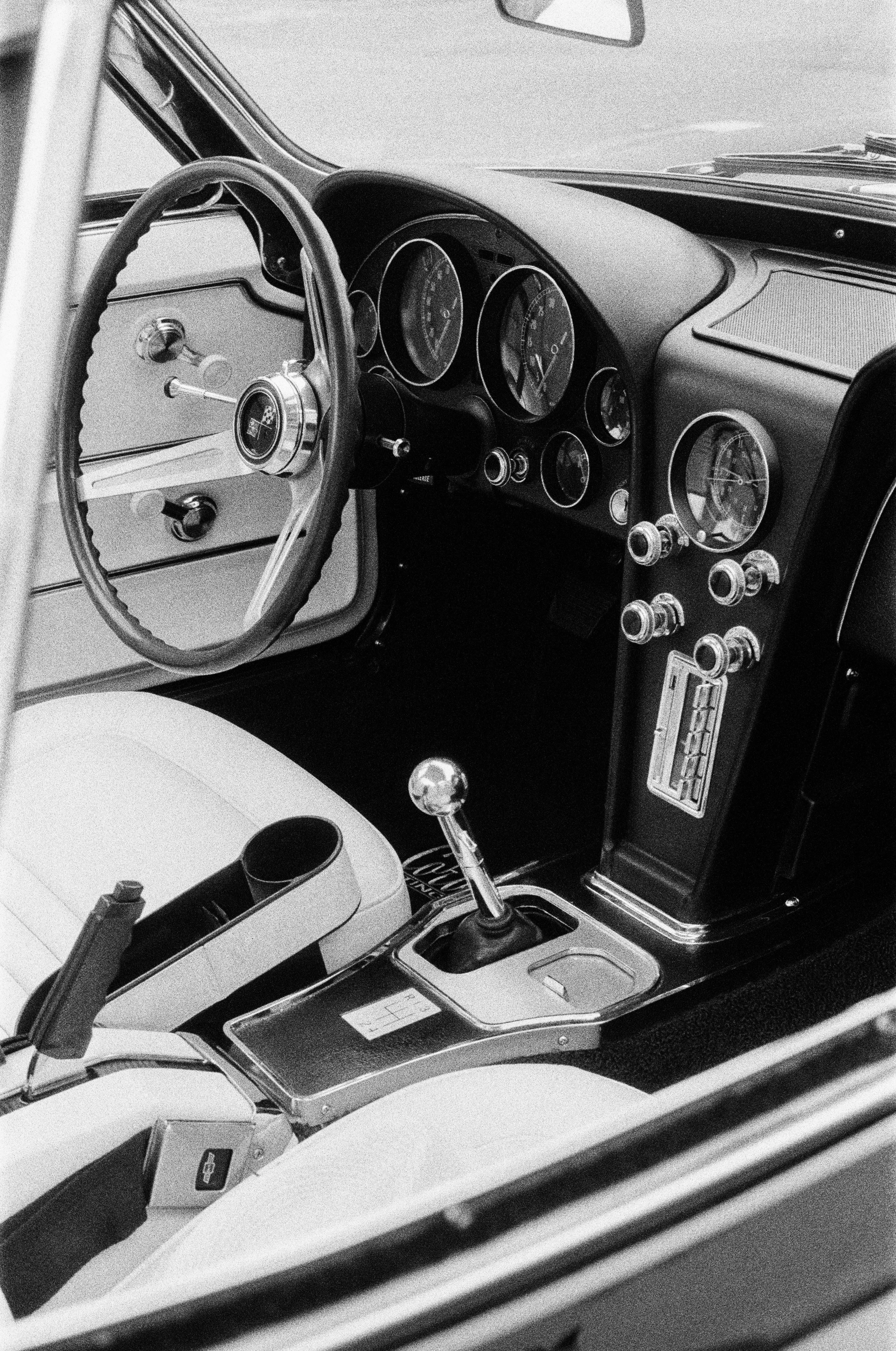 Black and white photo of a vintage Corvette car's interior dashboard with gauges, controls, and a gear shift lever. Photo by Vincent Prograce