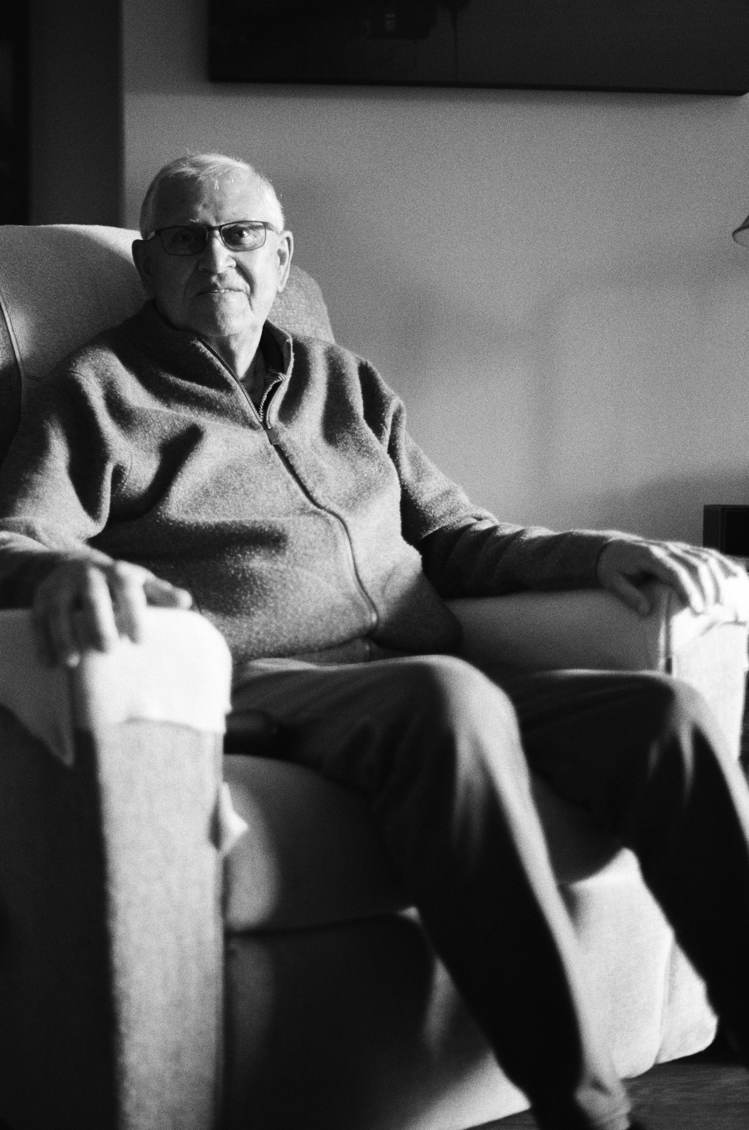 An elderly man sitting in a cushioned armchair, wearing glasses and a zip-up sweater, looking at the camera in a home setting, depicted in black and white. Photo by Vincent Prograce