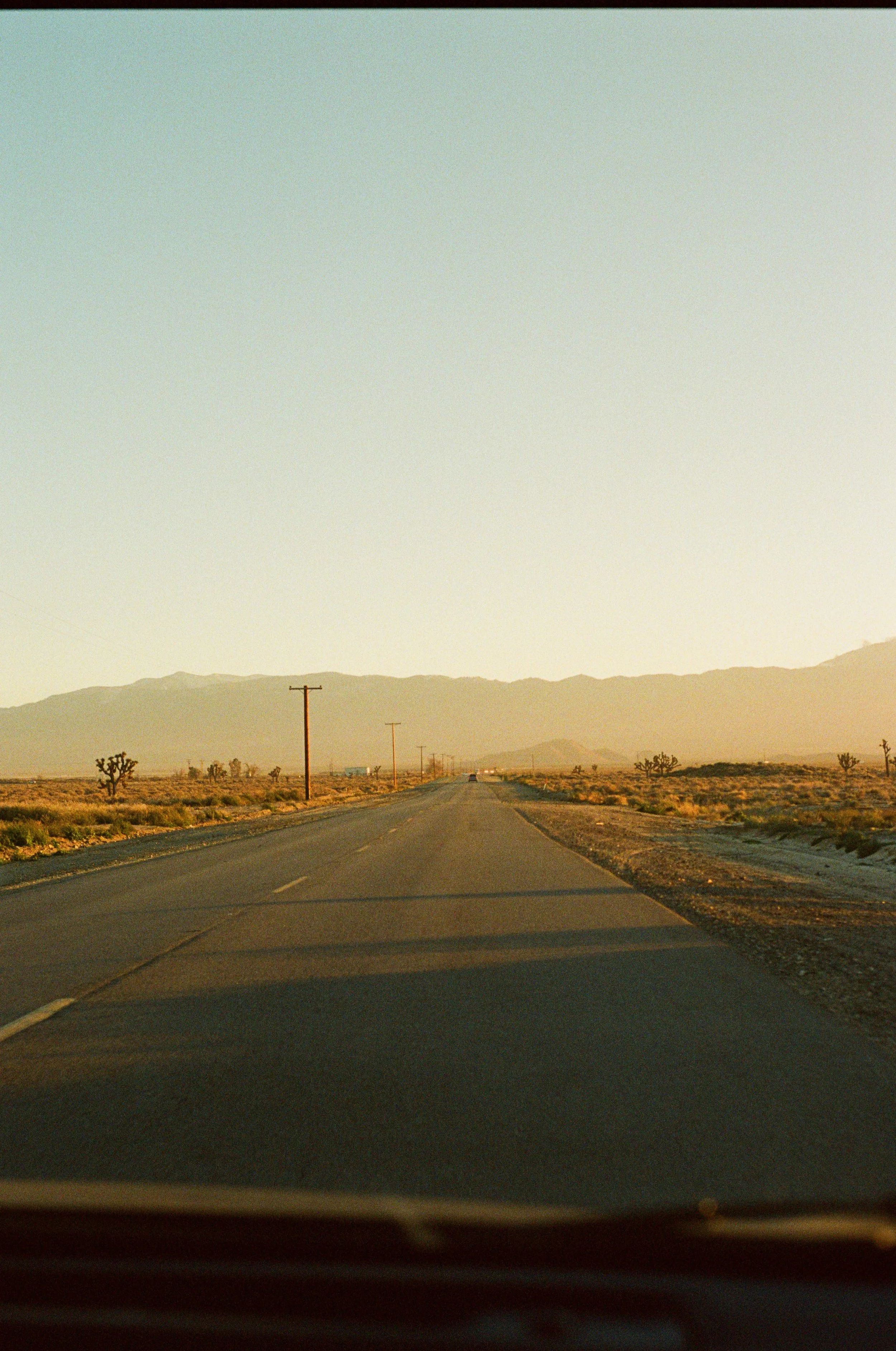 A straight desert highway with sparse vegetation and mountains in the distance, taken from inside a moving vehicle during sunset or sunrise. Photo by Vincent Prograce