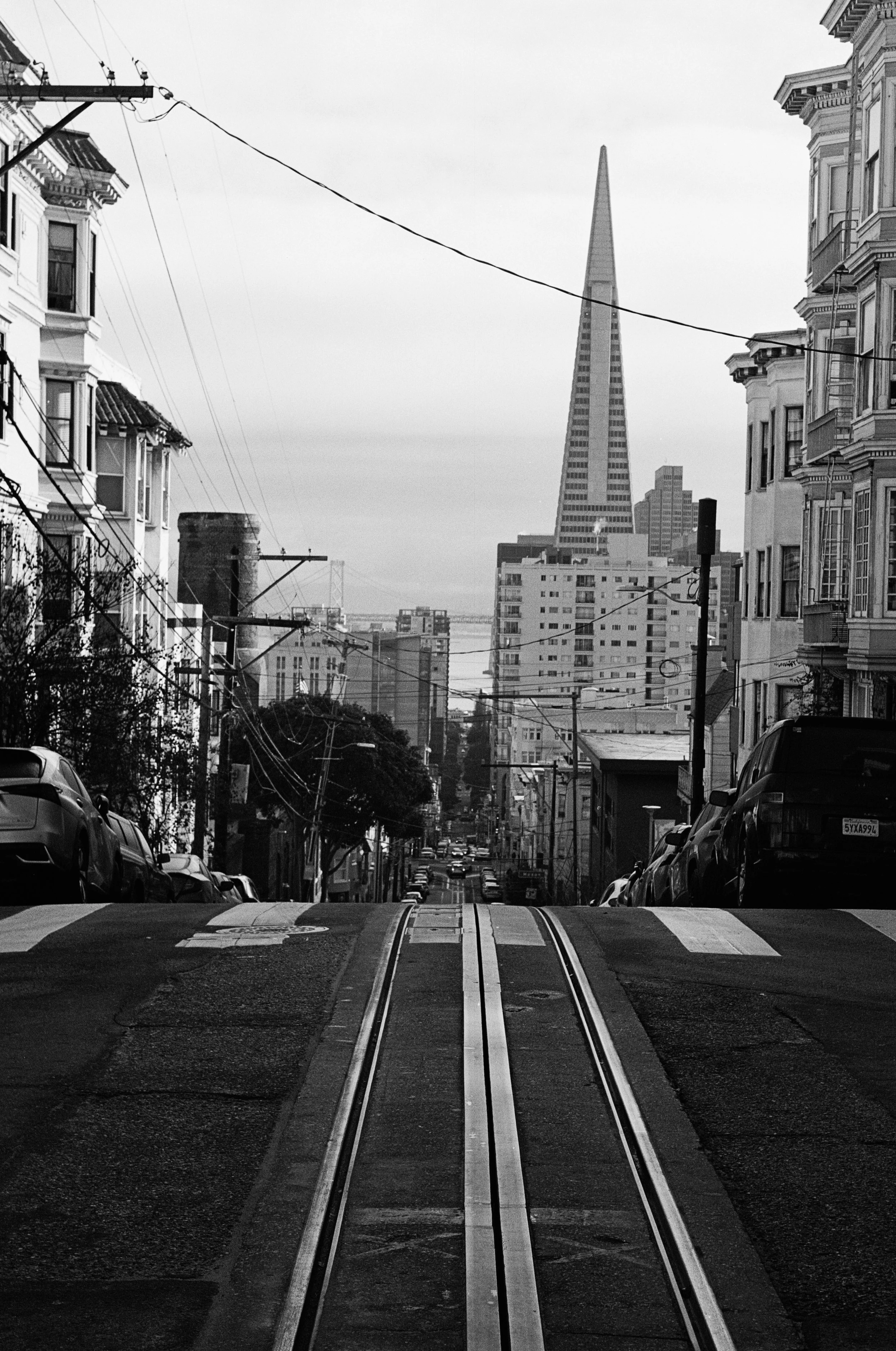 Black and white photo of a city street with cable car tracks and cars parked along the sides, leading towards the Transamerica Pyramid building in San Francisco. Photo by Vincent Prograce