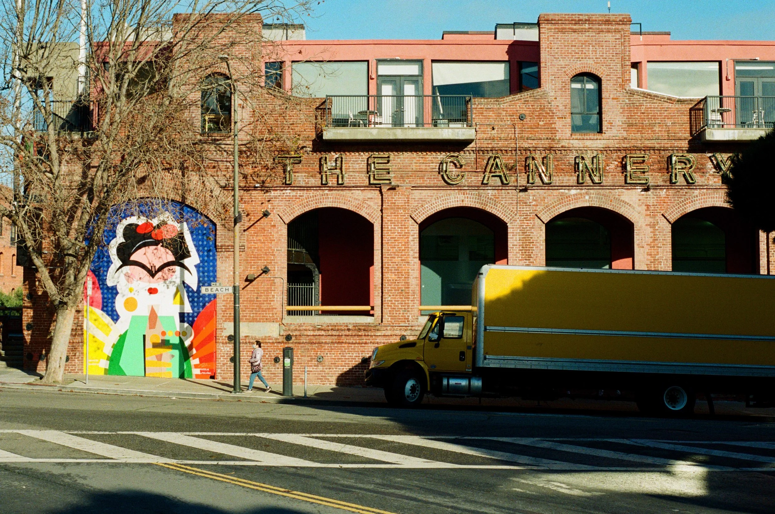 A brick building with large arched openings and a sign that reads 'The Cannery'. A colorful mural of a stylized woman with sunglasses, earrings, and a red bow. A yellow truck parked in front, with a person walking on the sidewalk nearby. Photo by Vin