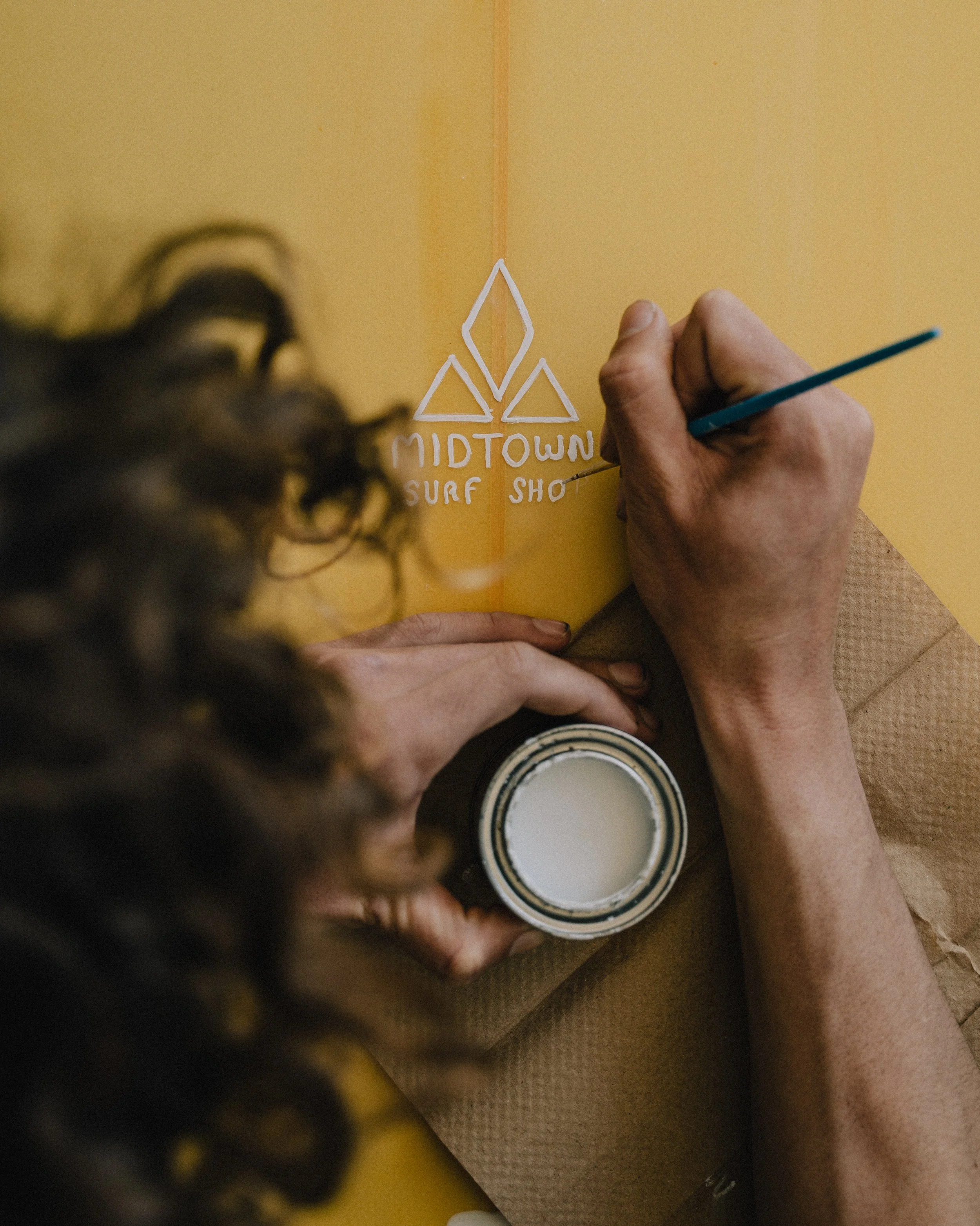 A person is painting a logo for 'Midtown Surf Shop' on a yellow surface, using a small brush and white paint. Photo by Vincent Prograce