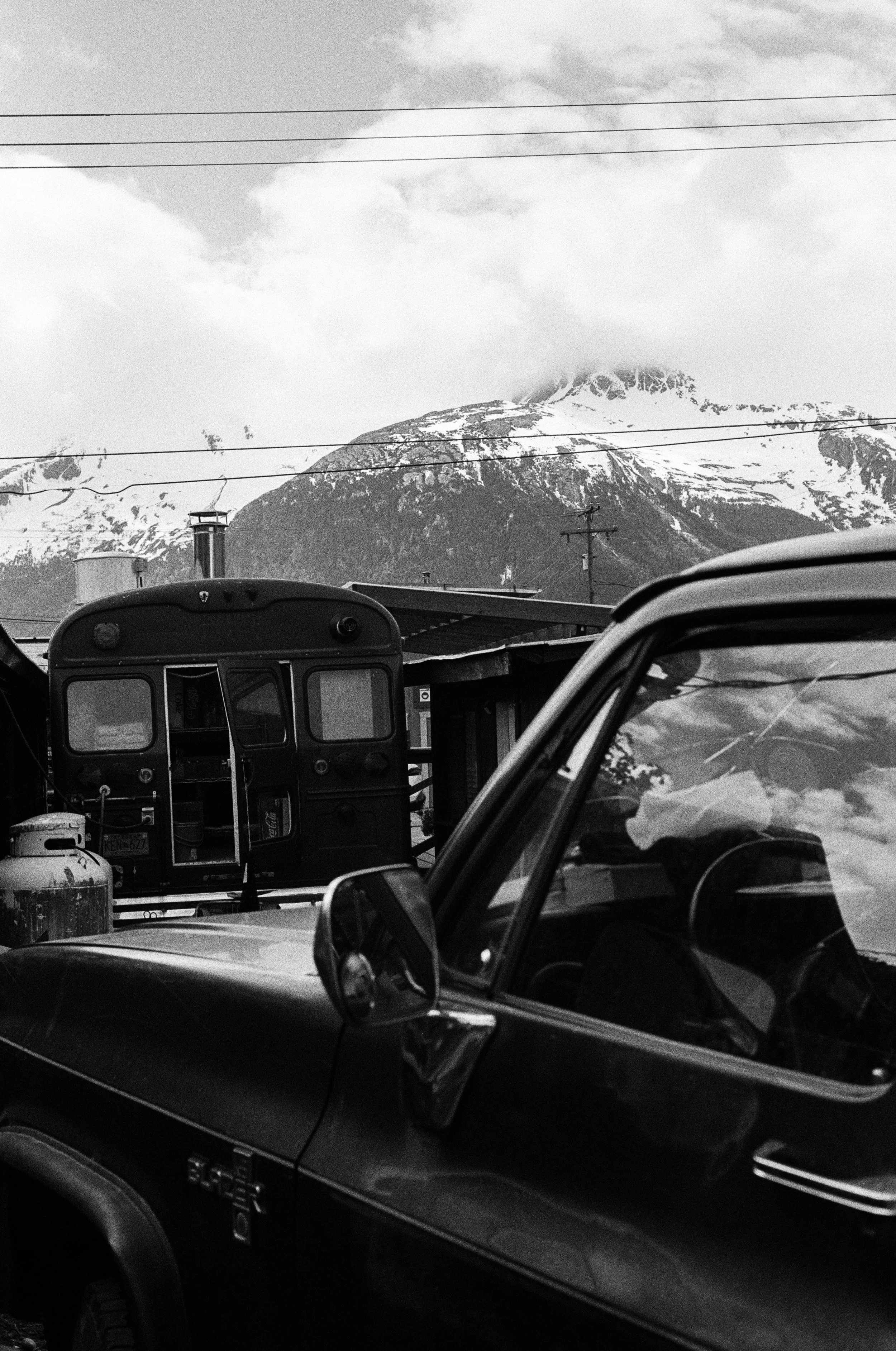 Black and white photo of a vintage vehicle and a small train in front of snow-capped mountains and cloudy sky. Photo by Vincent Prograce
