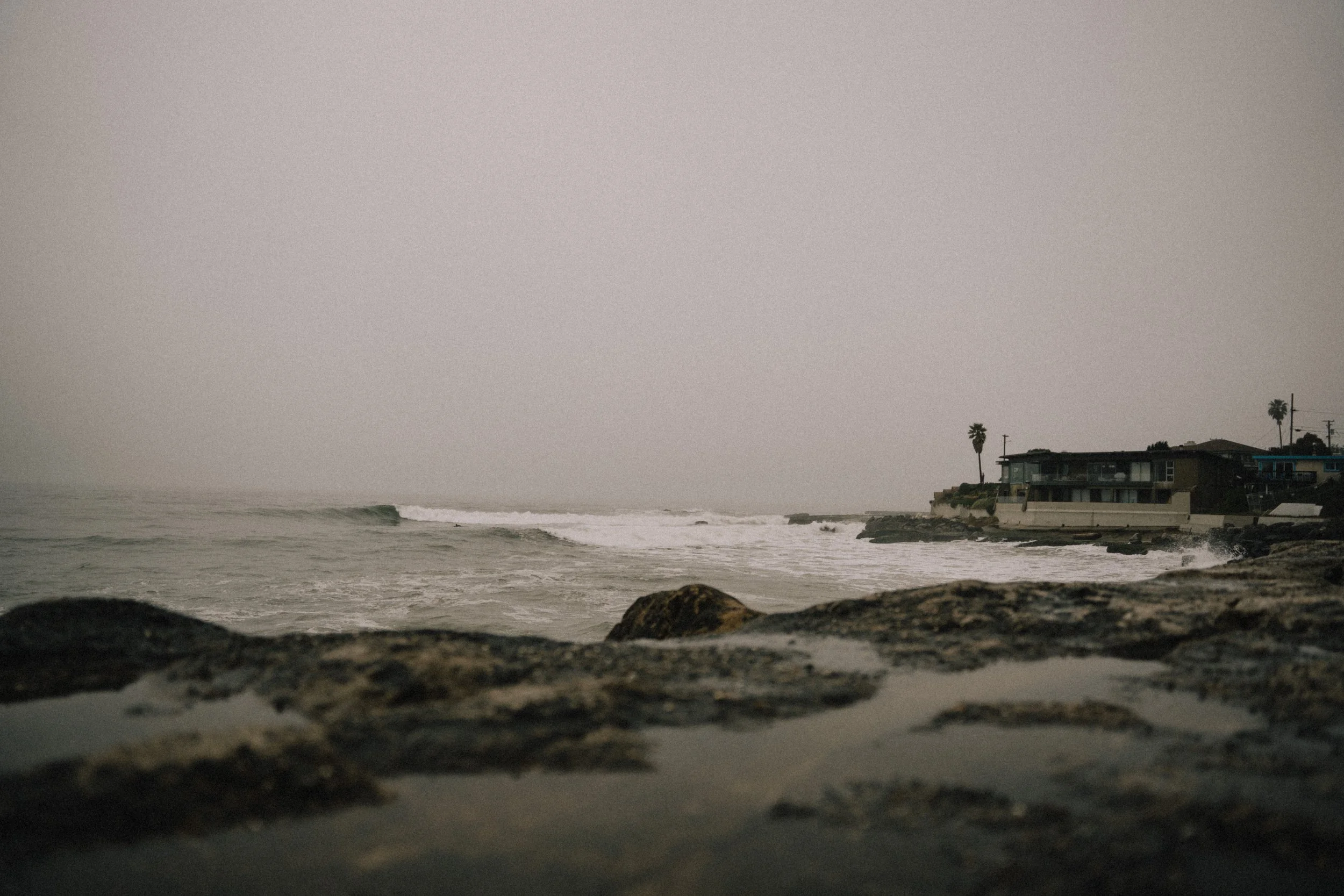 Overcast beach scene with rocks in the foreground, ocean waves, and beachfront houses with palm trees in the background. Photo by Vincent Prograce