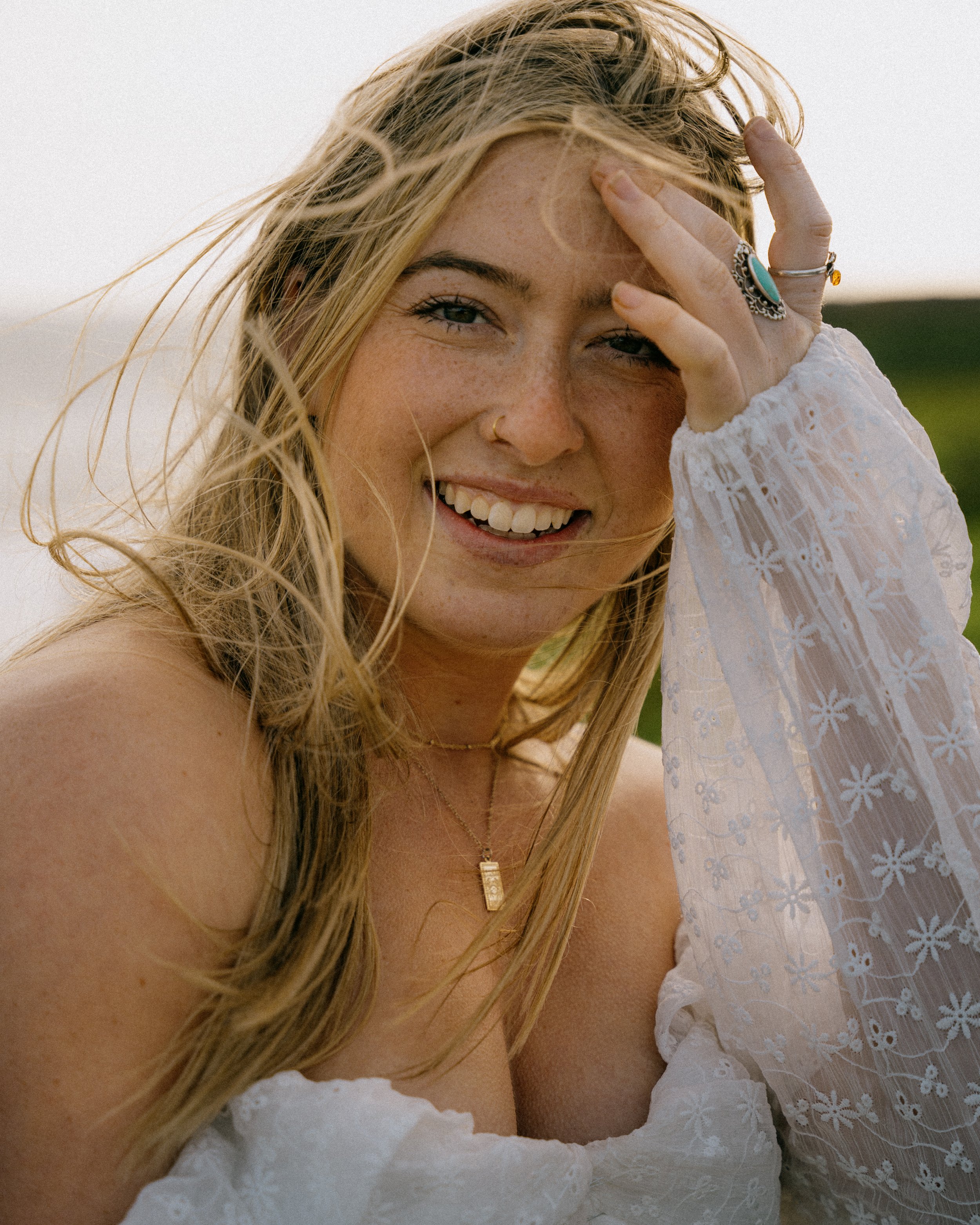 A smiling woman with blonde hair, wearing a white dress, jewelry, and a ring, outdoors on a windy day. Photo by Vincent Prograce