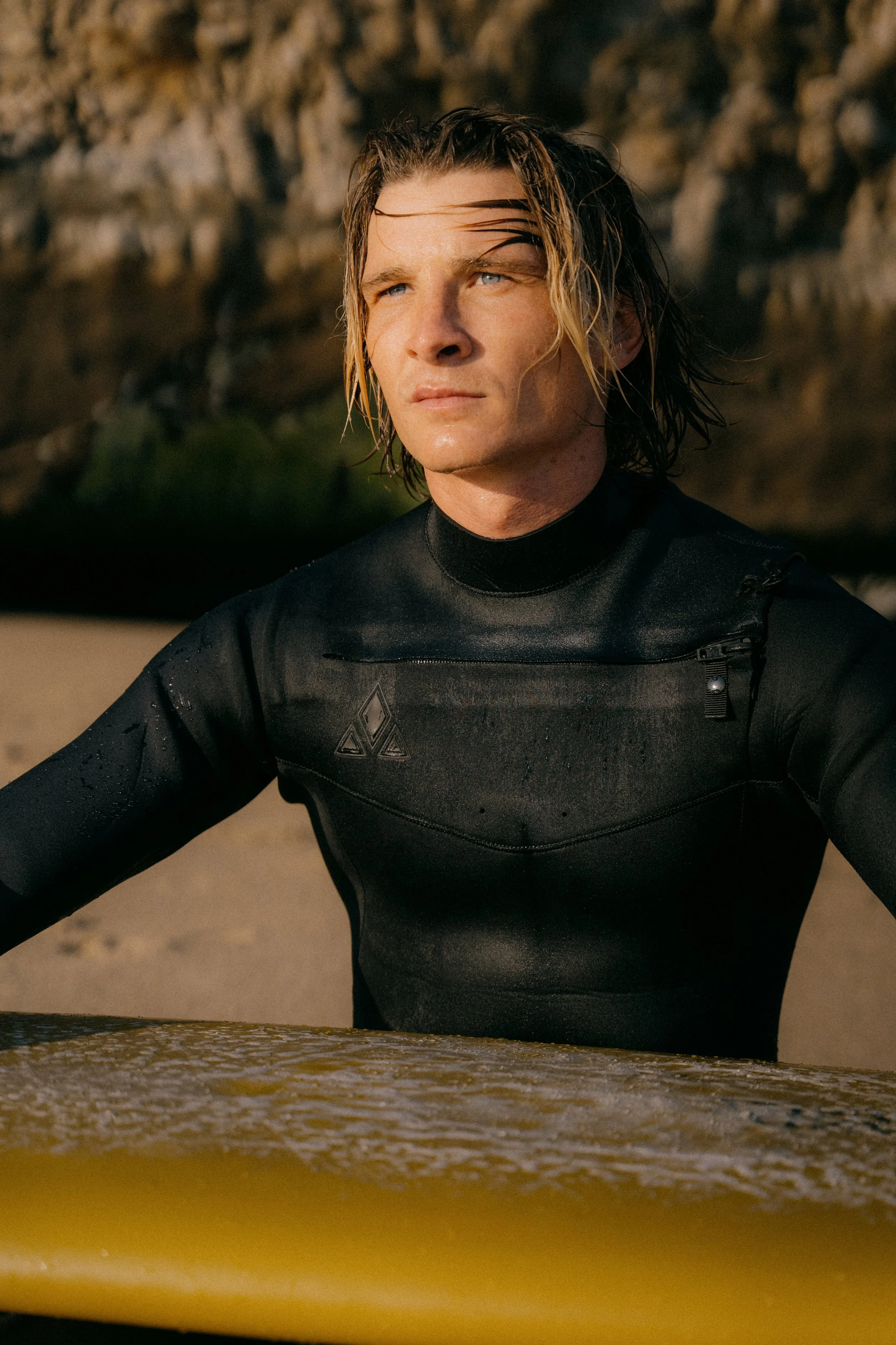 A man with wet hair, wearing a black wetsuit, sitting at the edge of a surfboard on a beach. Photo by Vincent Prograce