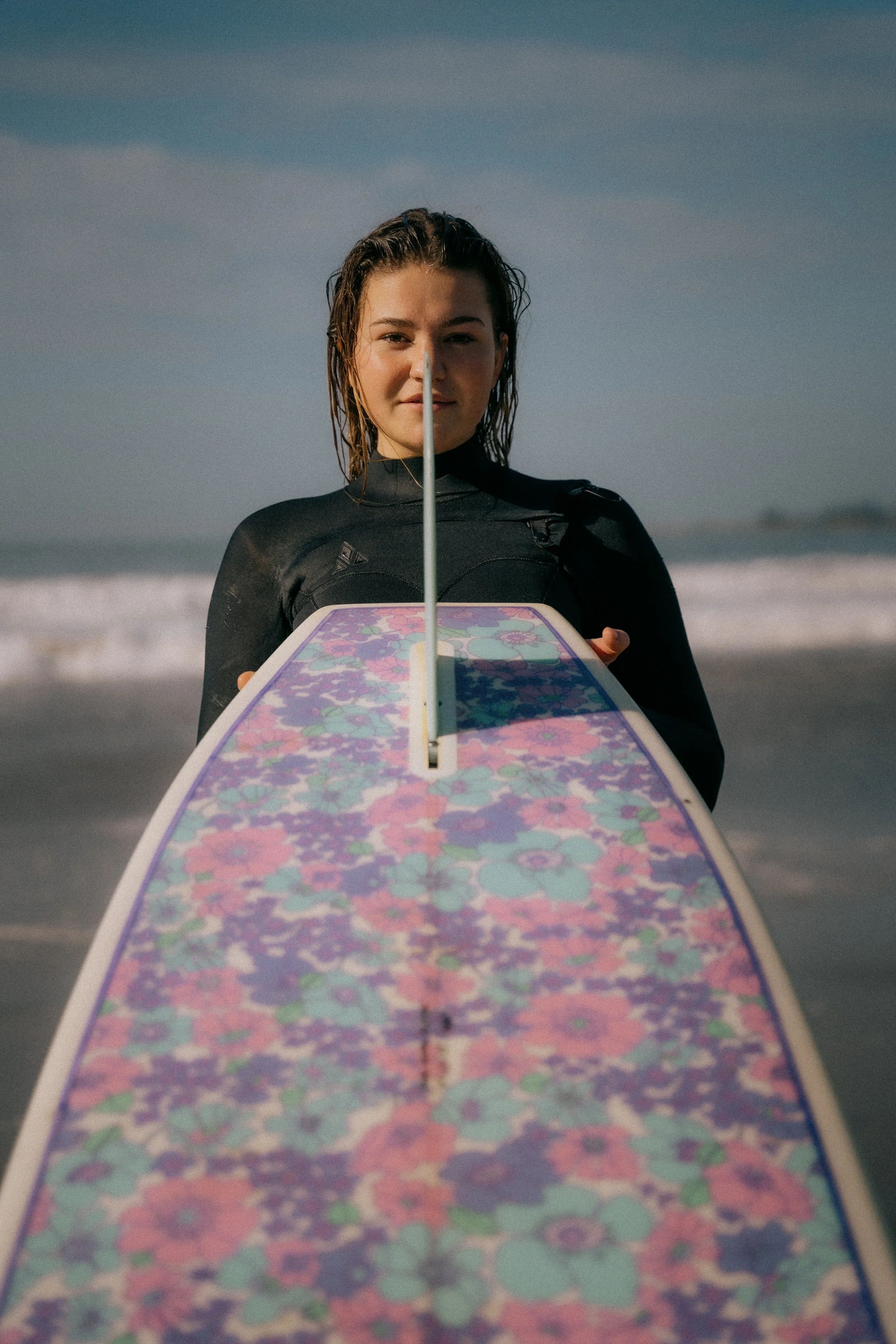 Young woman holding a surfboard with a colorful floral pattern on the beach, facing the camera with ocean waves in the background. Photo by Vincent Prograce