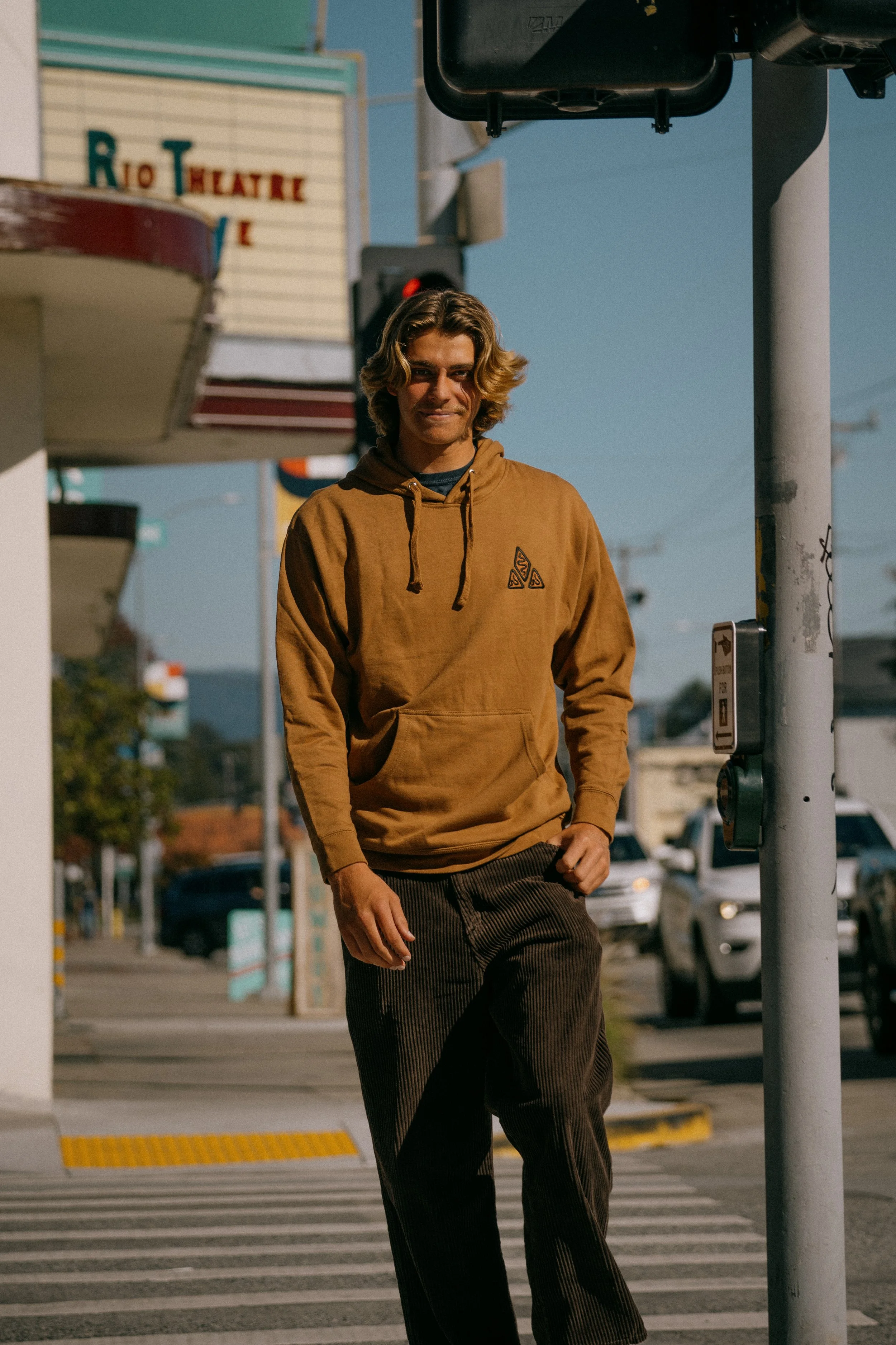 A person with wavy hair walking across a crosswalk on a sunny day, wearing a tan hoodie and brown pants, with a vintage theater sign in the background. Photo by Vincent Prograce