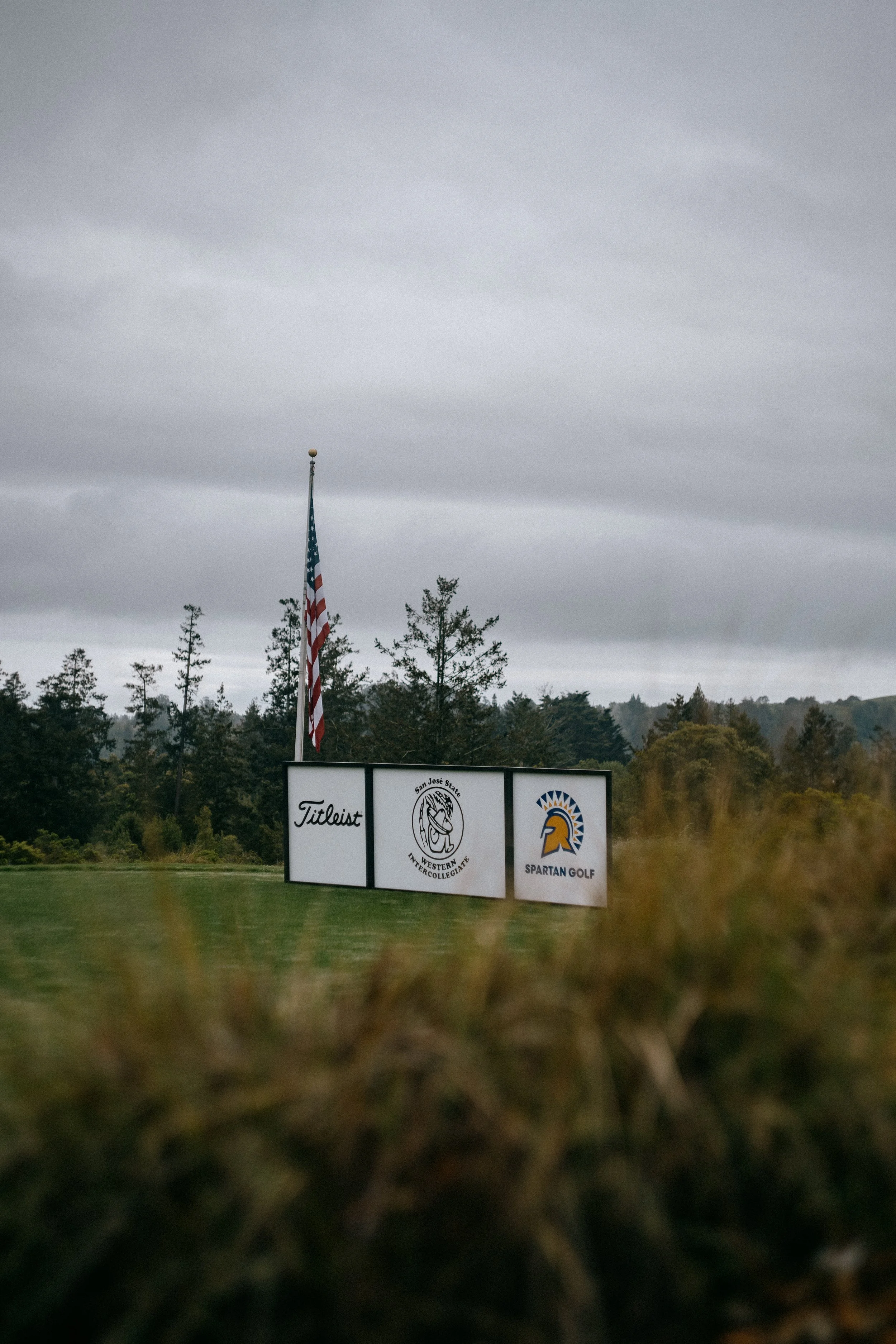 A golf course with a sign displaying the logos of Titleist, San Jose State University, and Spartan Golf under an overcast sky, with the American flag nearby surrounded by trees. Photo by Vincent Prograce